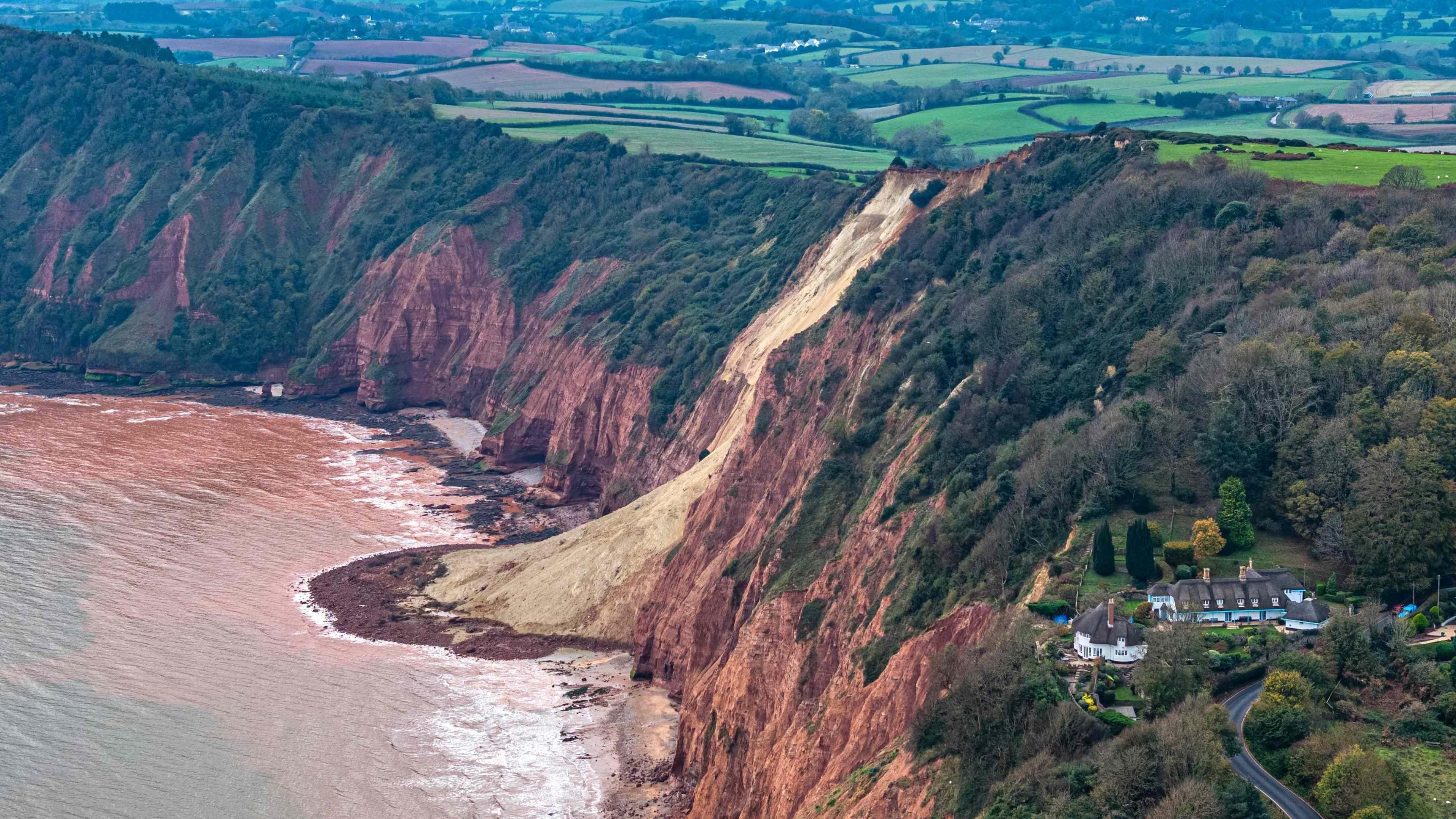 A house with a conical roof sits close to the edge above the sea with more houses further inland and nearby hedges and trees and a road along with signs of a cliff fall nearby with mud going into the sea.