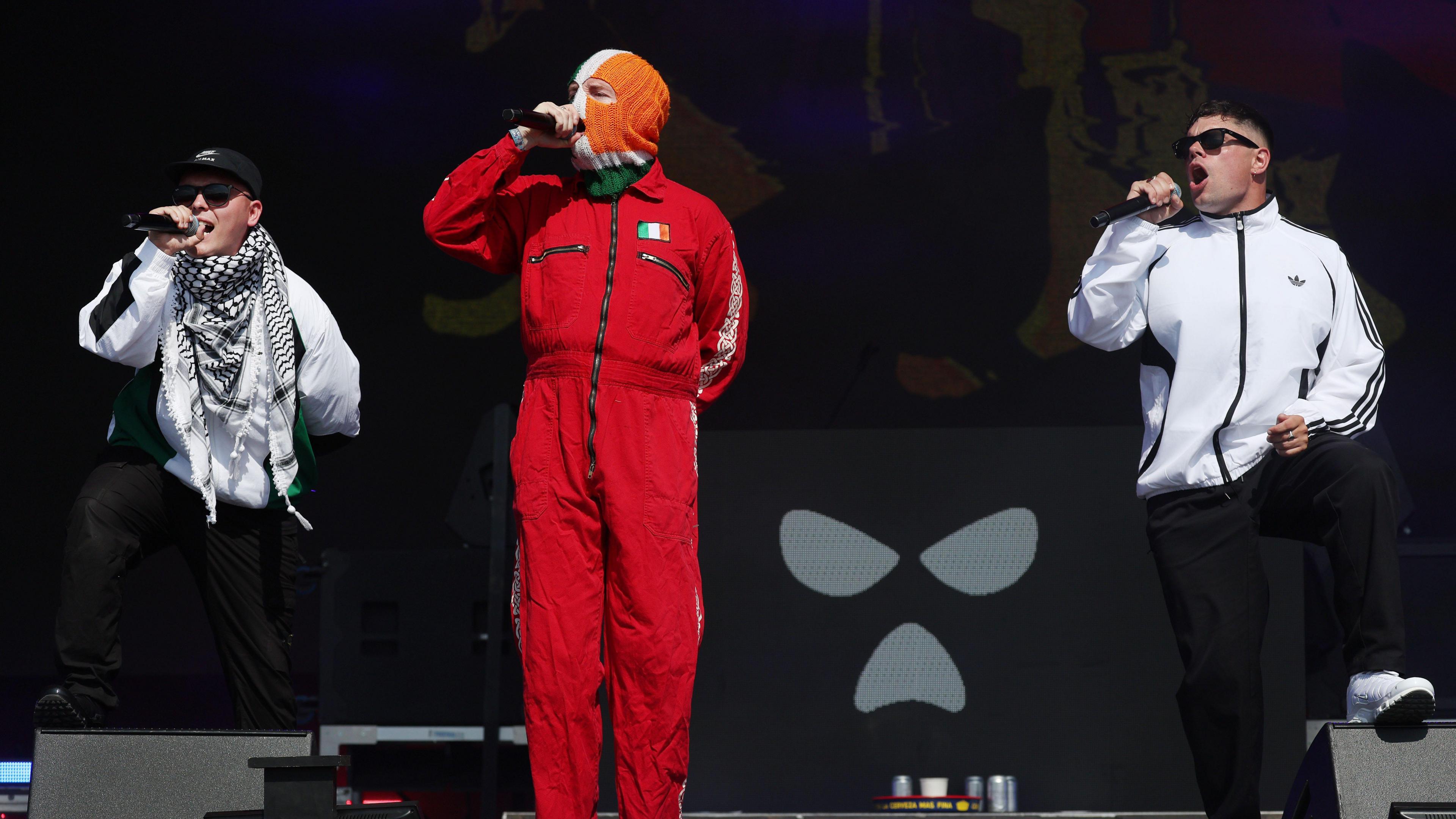 The band Kneecap perform onstage - all three members of the band are holding microphones. One of the group, standing centre, is wearing all red with a small Irish flag printed on it and a tricolour balaclava. The other two band members are wearing white jackets and sunglasses.