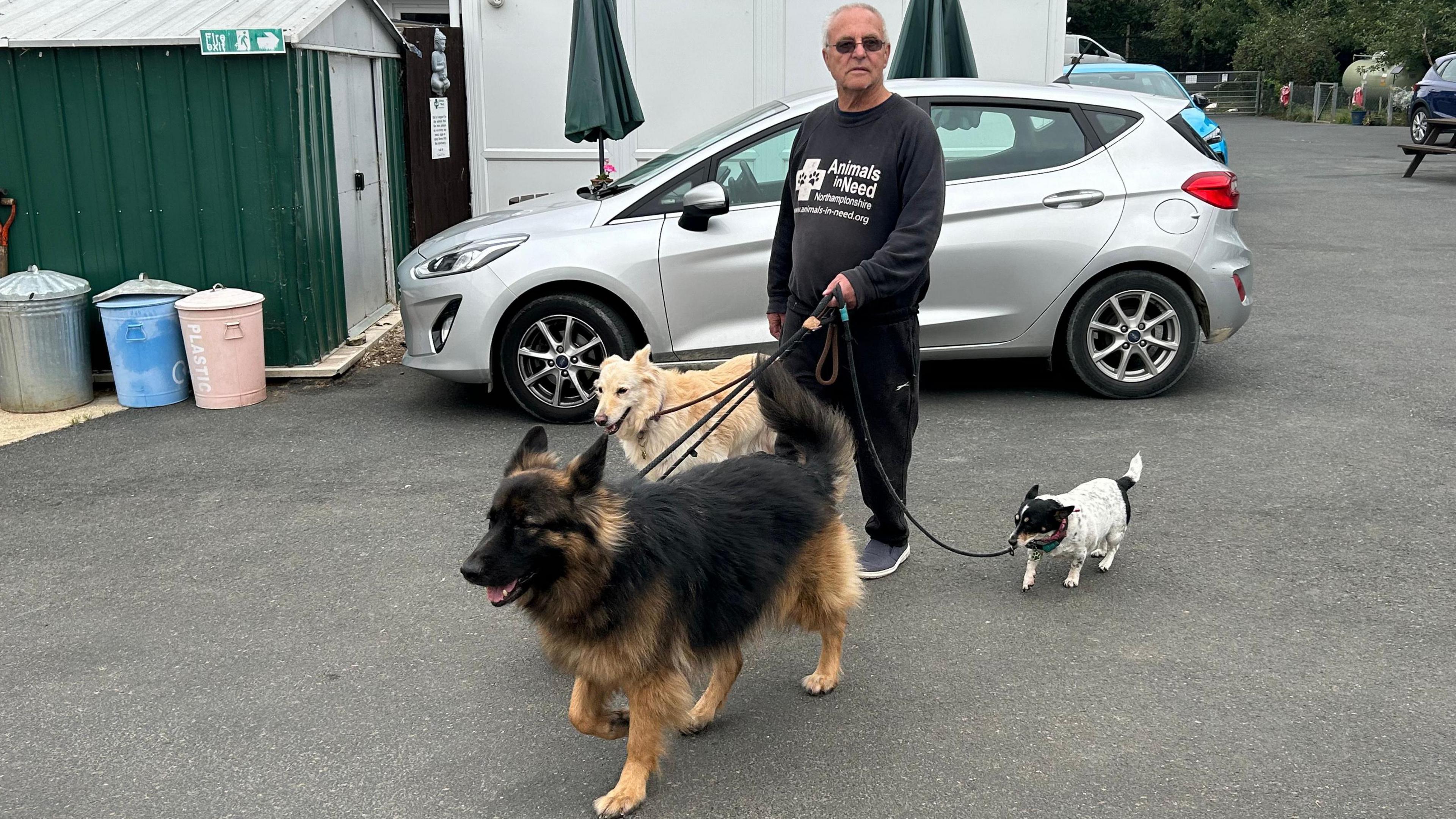 Roy Marriott wearing a dark sweatshirt walking three dogs in a carpark with a silver car behind him