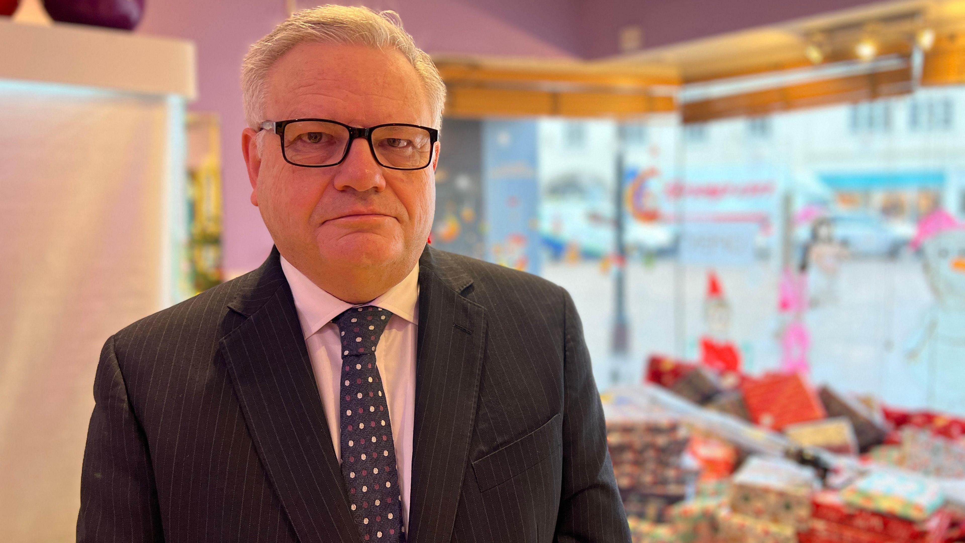 The Minister looks at the camera with the presents in the background in the cafe. He is wearing a black jacket with thin white strips and a blue dotted tie. He has a pink shirt on with black rectangular and has white hair.