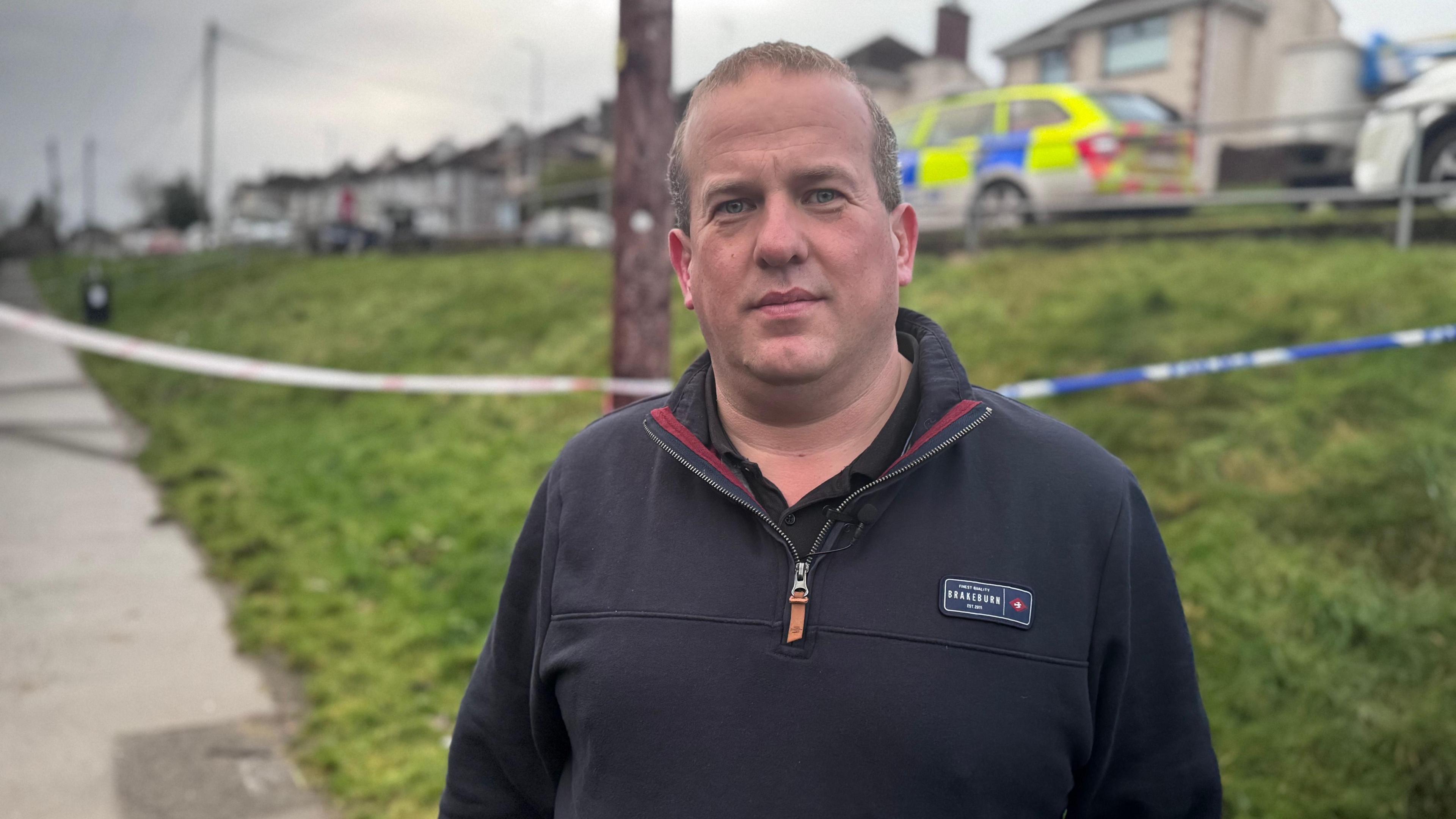 Clement Cuthebertson has short fair hair, and is wearing a quarter zip pullover. Behind him is a grass bank, and beyond, a police car parked on the road. 