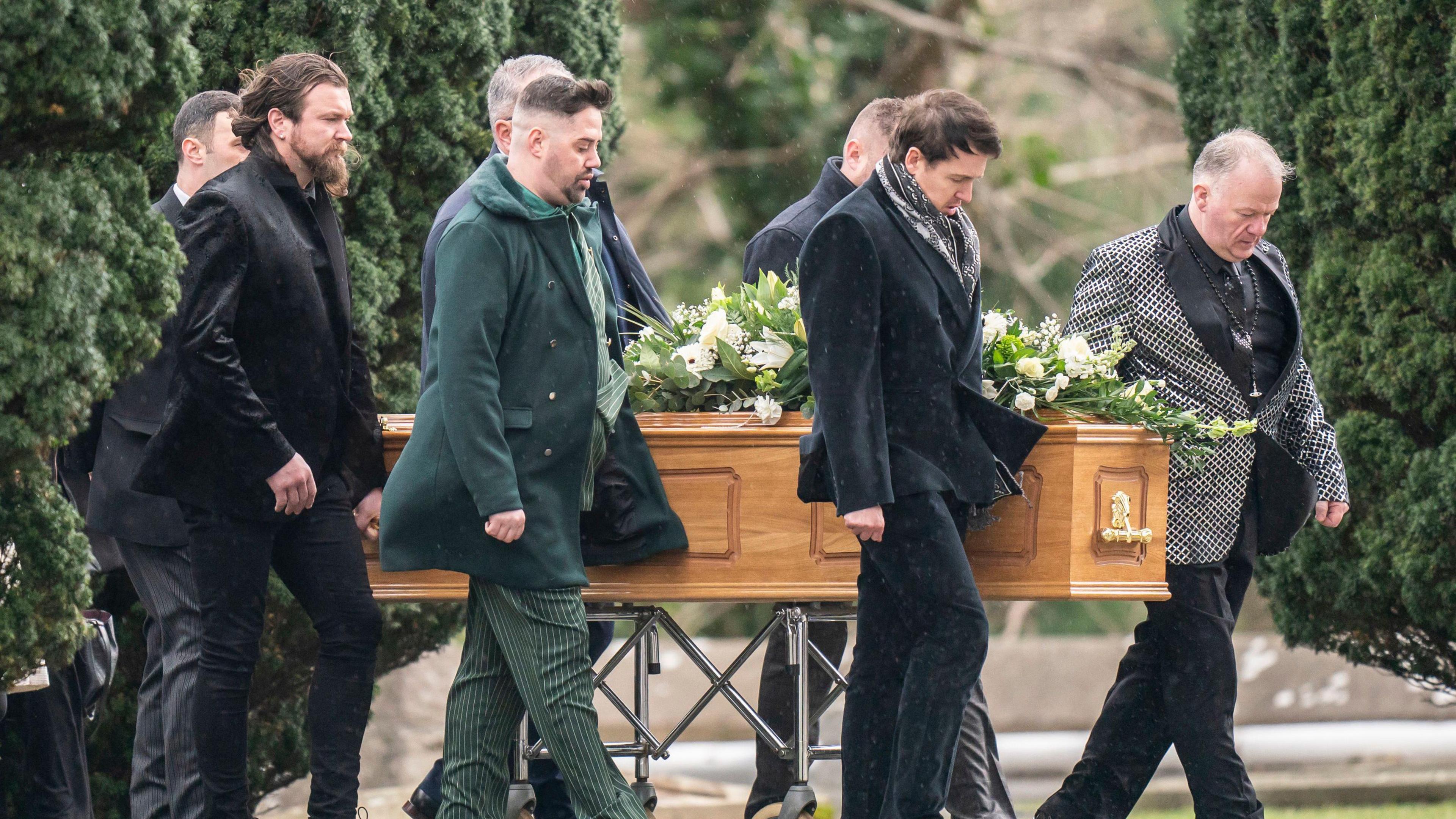 Seven men with their heads bowed walk alongside a light-brown wooden coffin, which is supported by a metal trolley, against a backdrop of a church-yard.