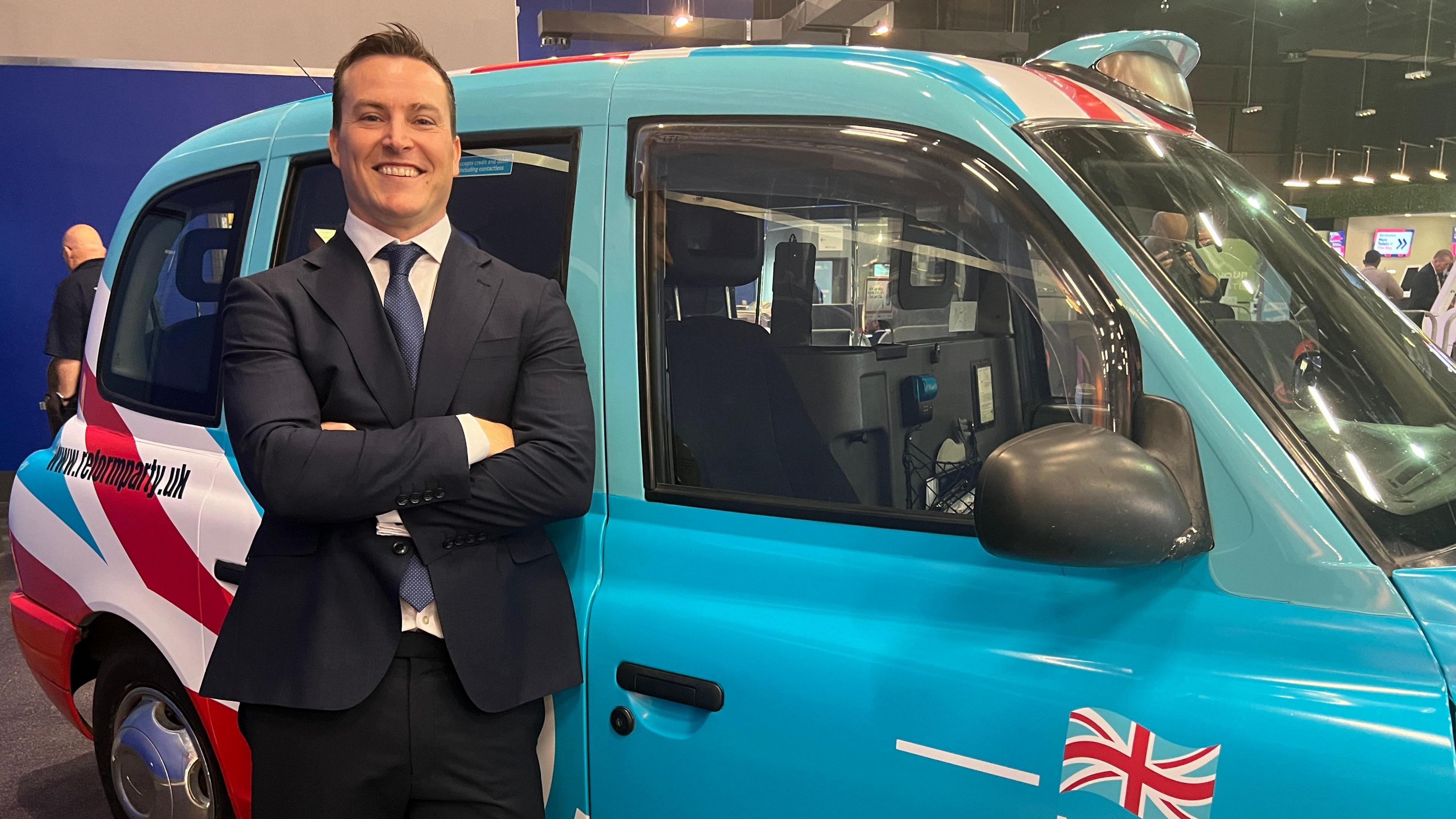 James McMurdock MP. He has brown hair and is smiling. He is standing in front of a light blue taxi with Union Jack flags on it. 