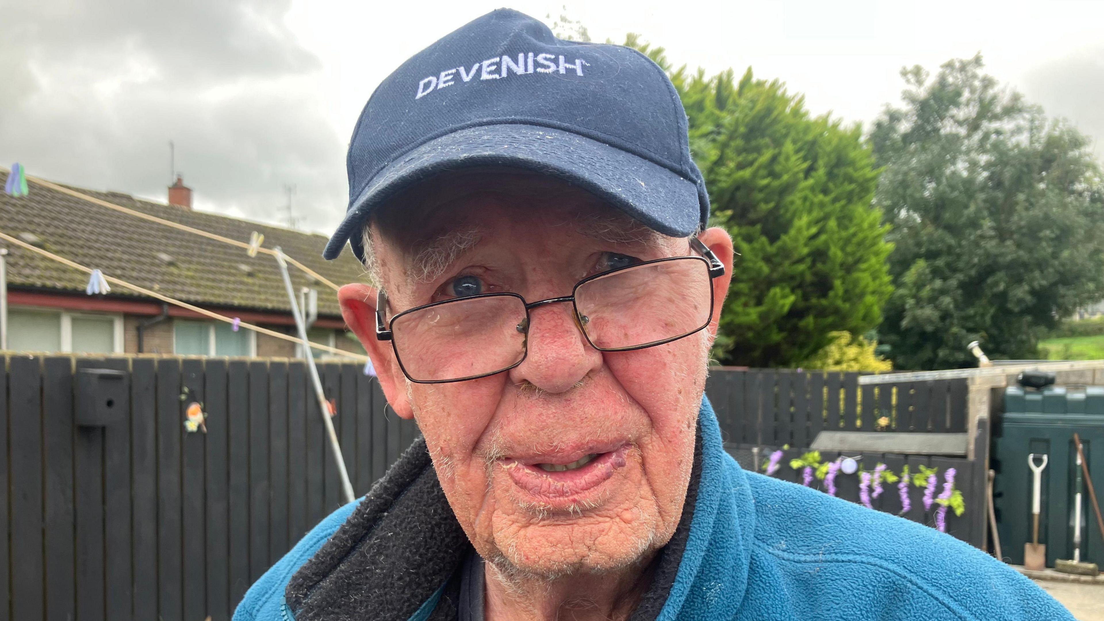 An elderly man with a grey hat with white writing in caps saying 'DEVENISH'. He is wearing a blue fleece and grey thin rimmed glasses and standing in a garden with grey painted wooden walls and a shovel in view.
