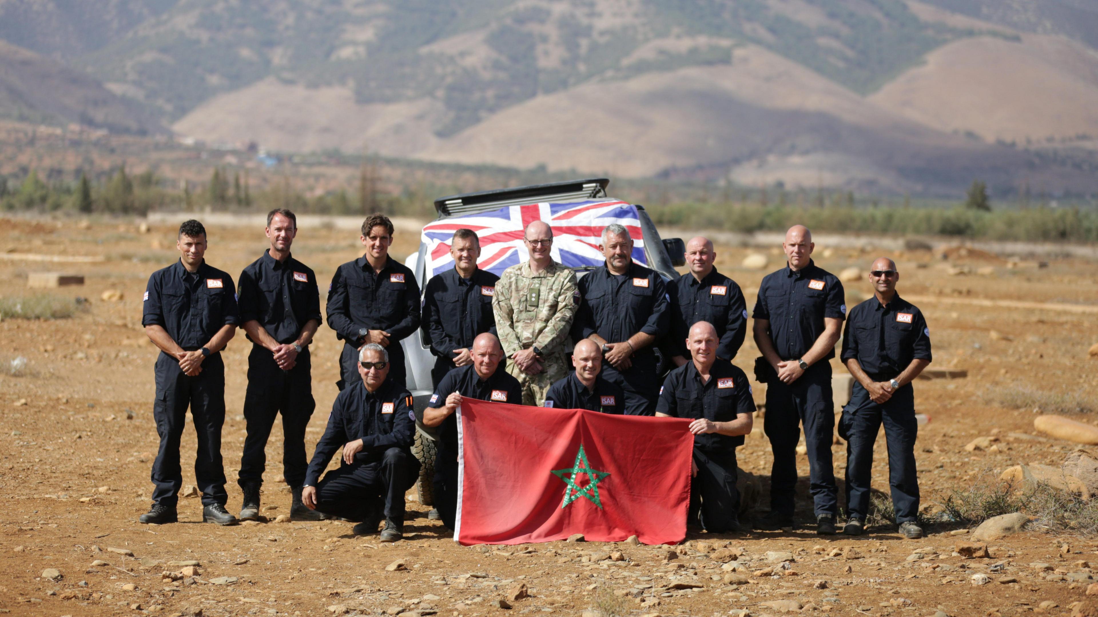 A group of men wearing black stand on sandy ground and hold a red flag with a green star on it. A car with a Union Jack flag on it is behind them. Mountains can be seen in the background.