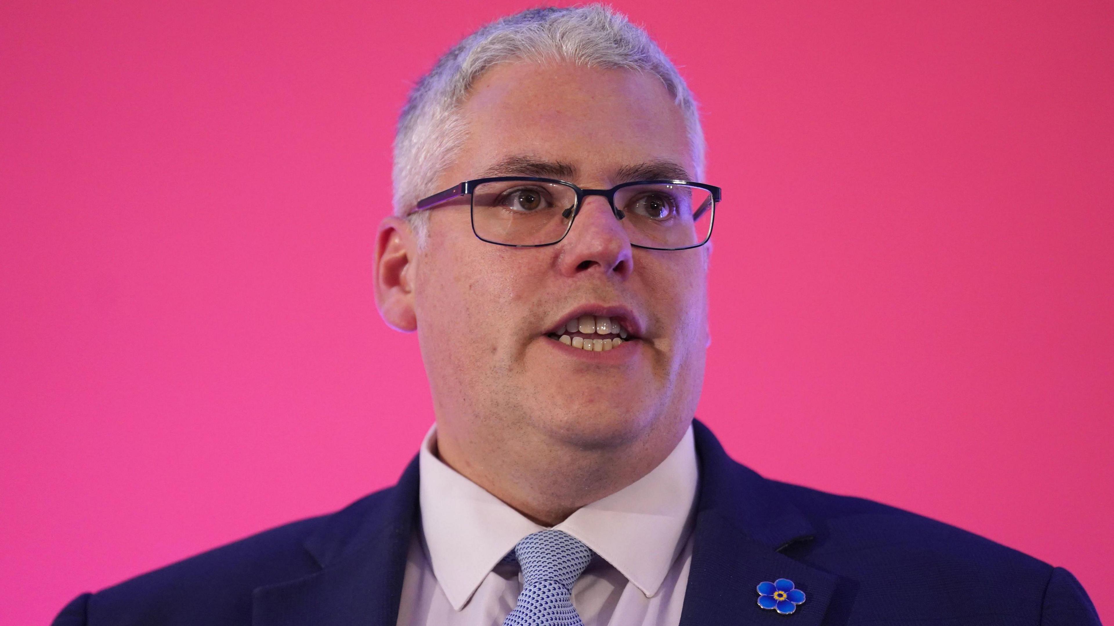 DUP leader Gavin Robinson stands in front of a pink screen. He has short grey hair, rectangle framed glasses. He is wearing a navy blazer, a white collared shirt and a blue and white spotted tie. He has a blue flower pin on his lapel.