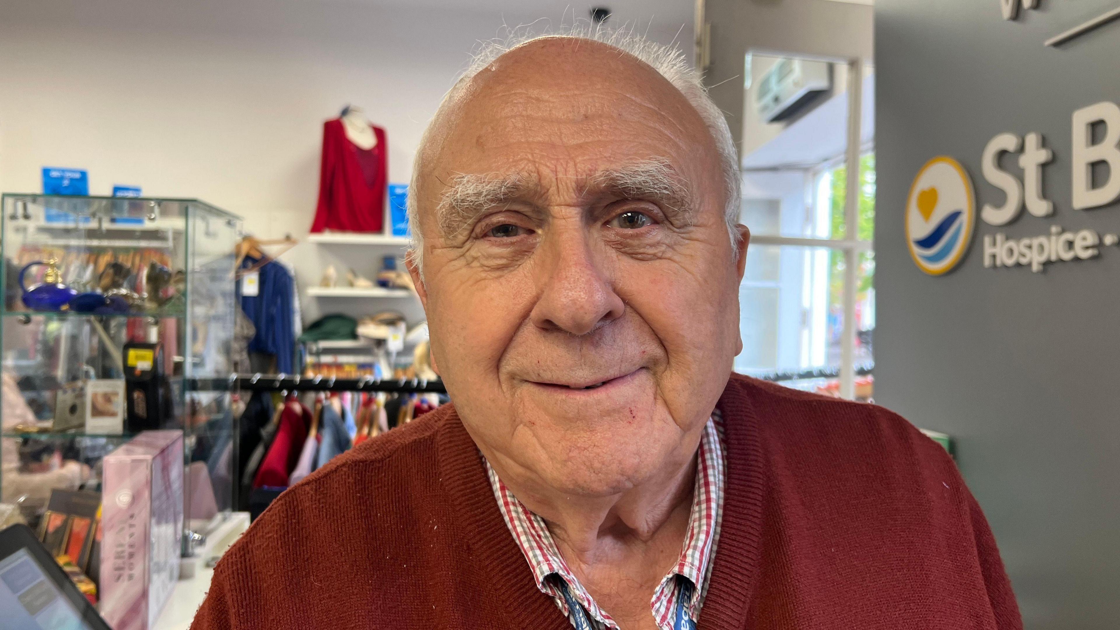 A man is stood in a charity shop smiling at the camera. He is wearing a red sweater and a red-and-white checked shirt. He has short grey hair.