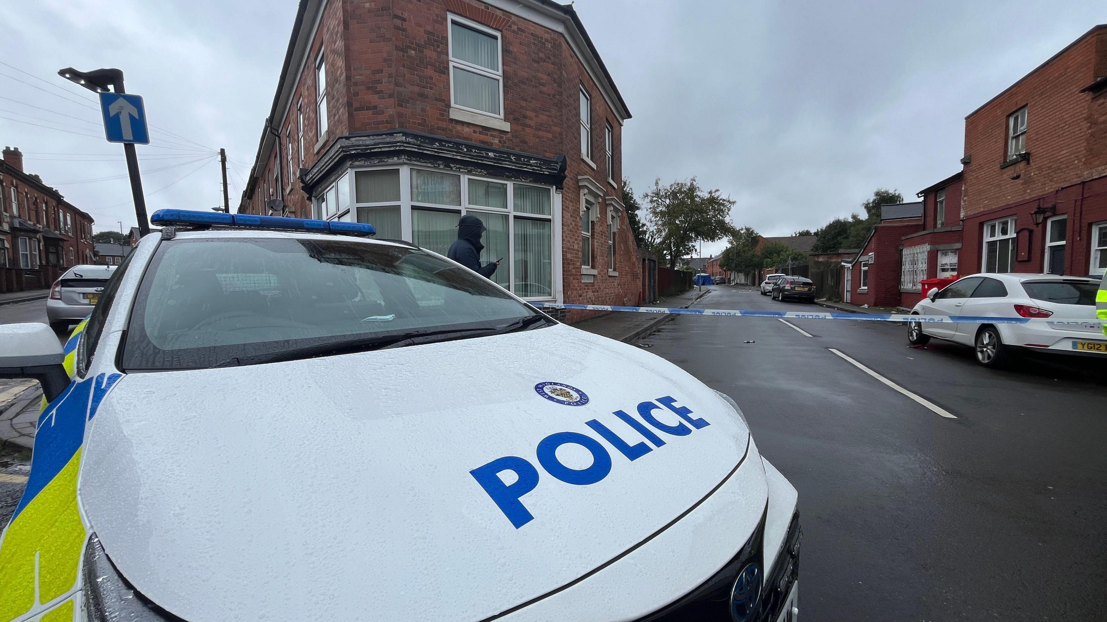 A police car next to a cordon, with blue and white police tape blocking the street. A blue police tent can be seen in the background.