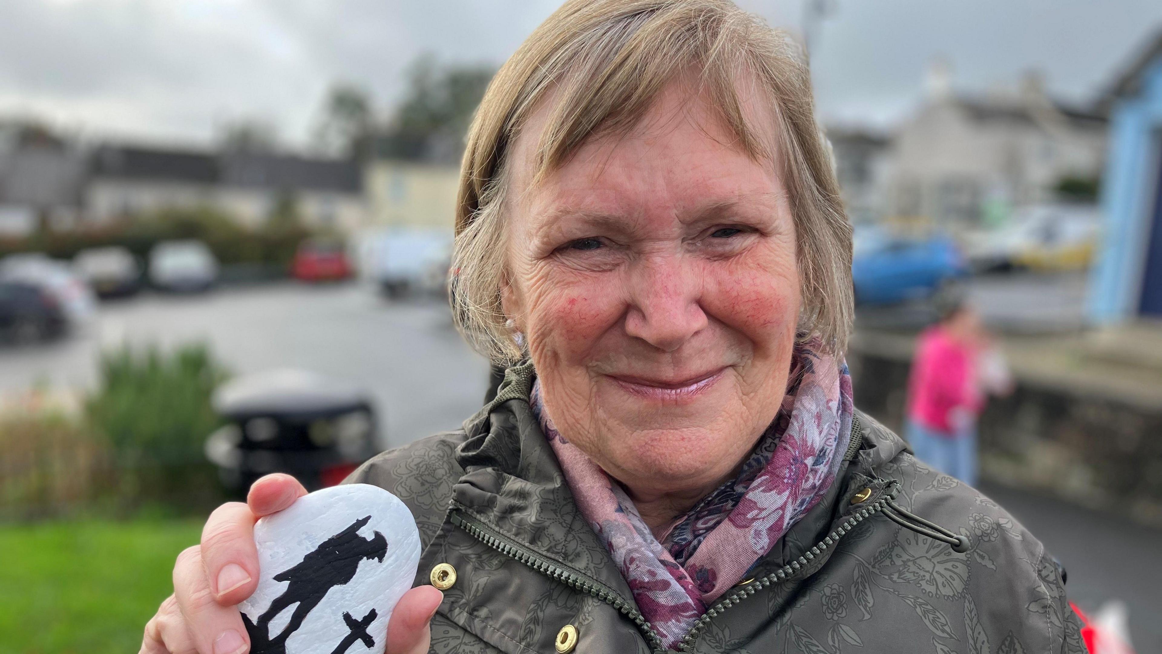 Woman with short mousey brown hair holding a pebble painted white with black silhouette of a soldier standing next to a grave. The woman is smiling at the camera and wearing a pink scarf around her neck and a green coat.