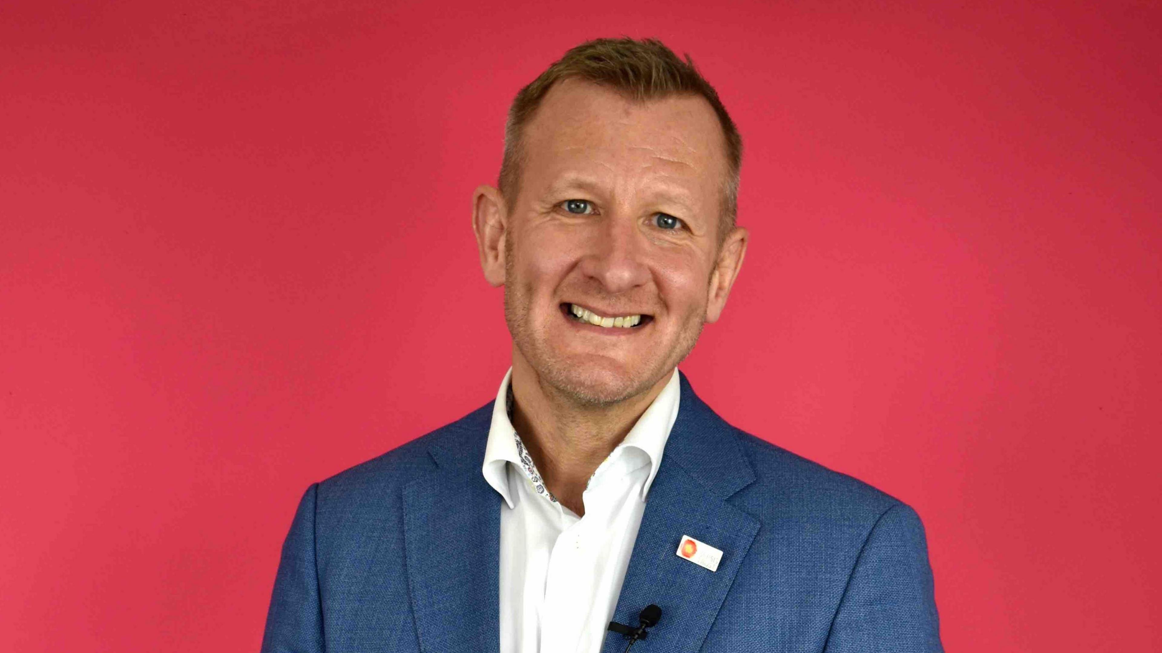 Ron Daniels wearing a blue blazer, white shirt and lapel badge with UK Sepsis Trust branding on it which can barely be made out. He has short, fair hair, and is looking at the camera. The background is entirely red.