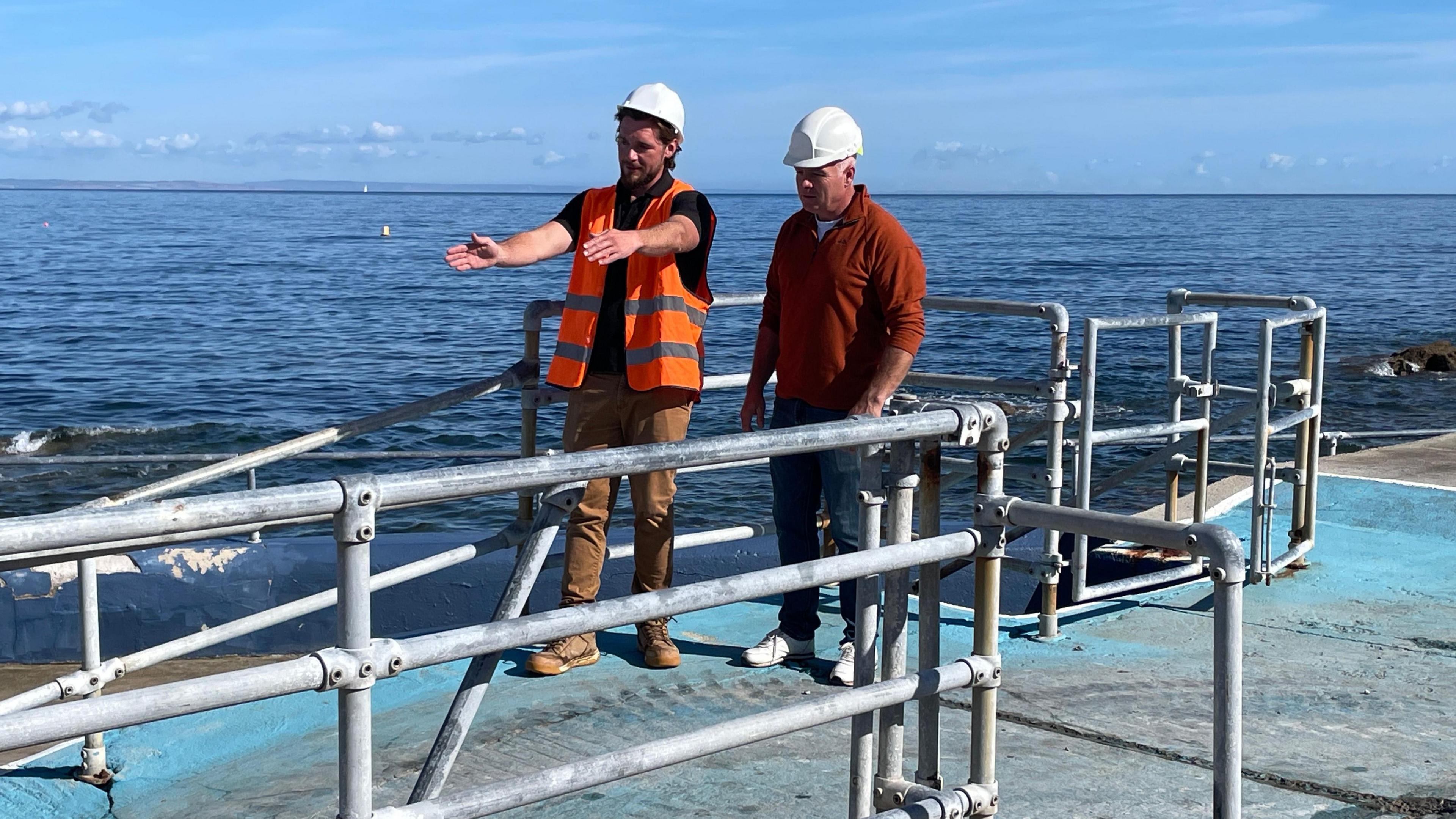 Two men wearing hard hats stand at the top of the ramp into the pool. The sea is being them.