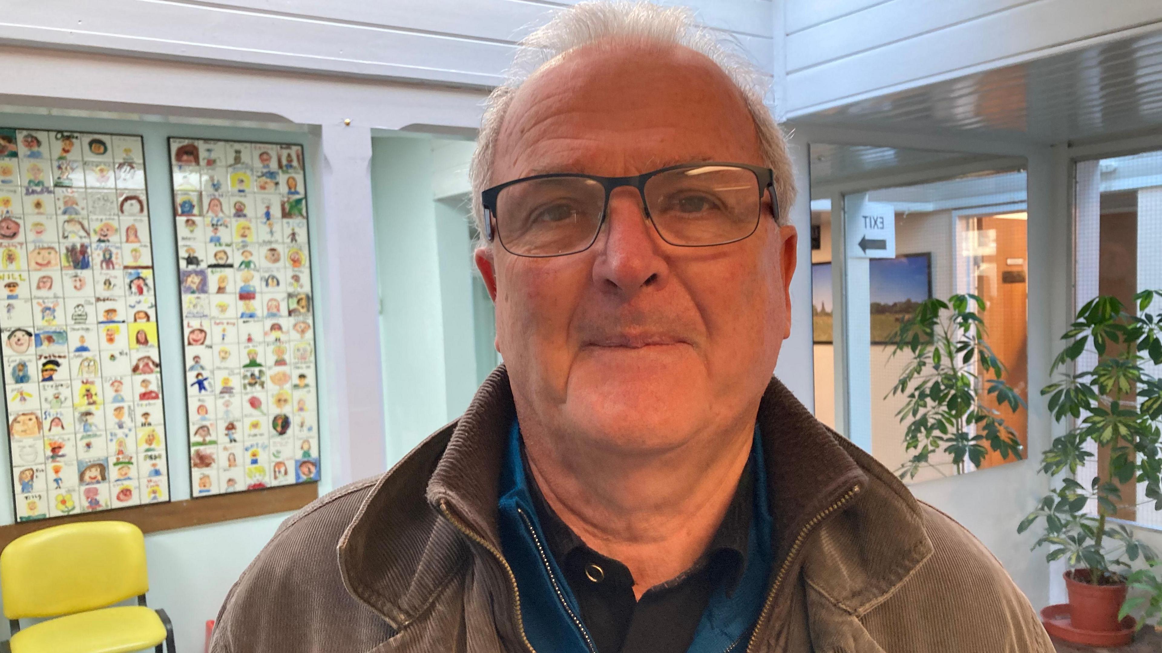 A man with grey hair, glasses, a black top and brown jacket looks at the camera. He is standing in the reception of a doctors surgery.