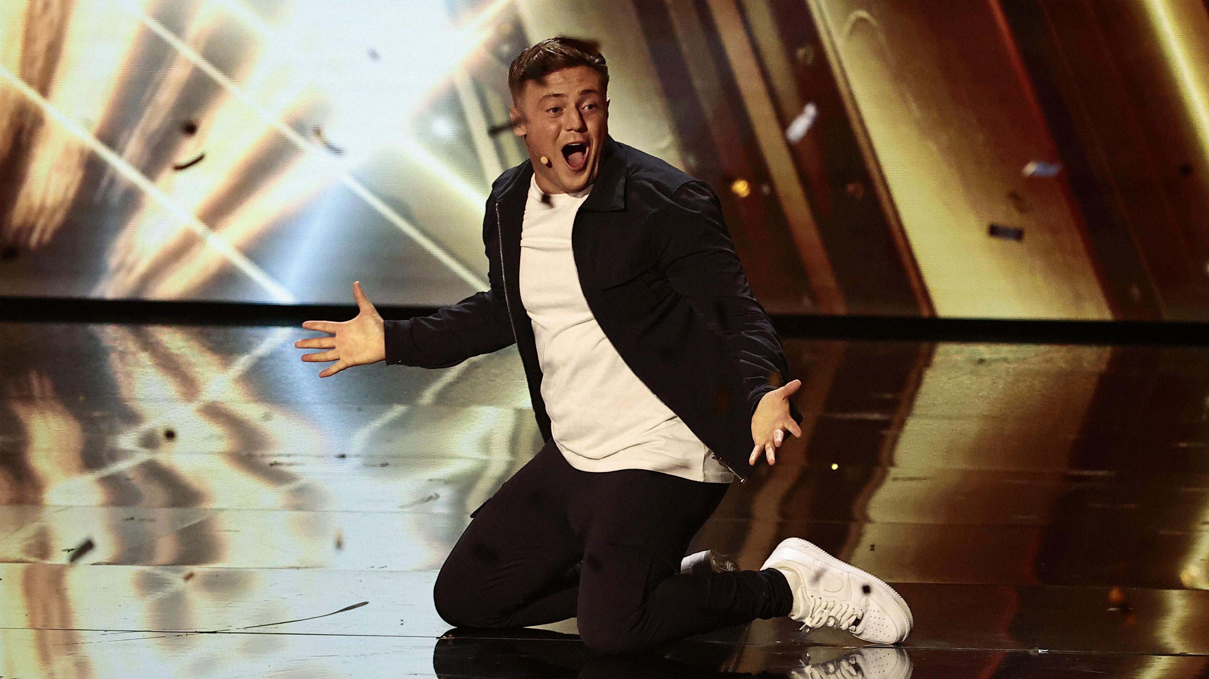 Harry Moulding on his knees with hands out on the stage as golden paper flies around. He is wearing black jeans and a black jacket with a white T-shirt and white trainers.