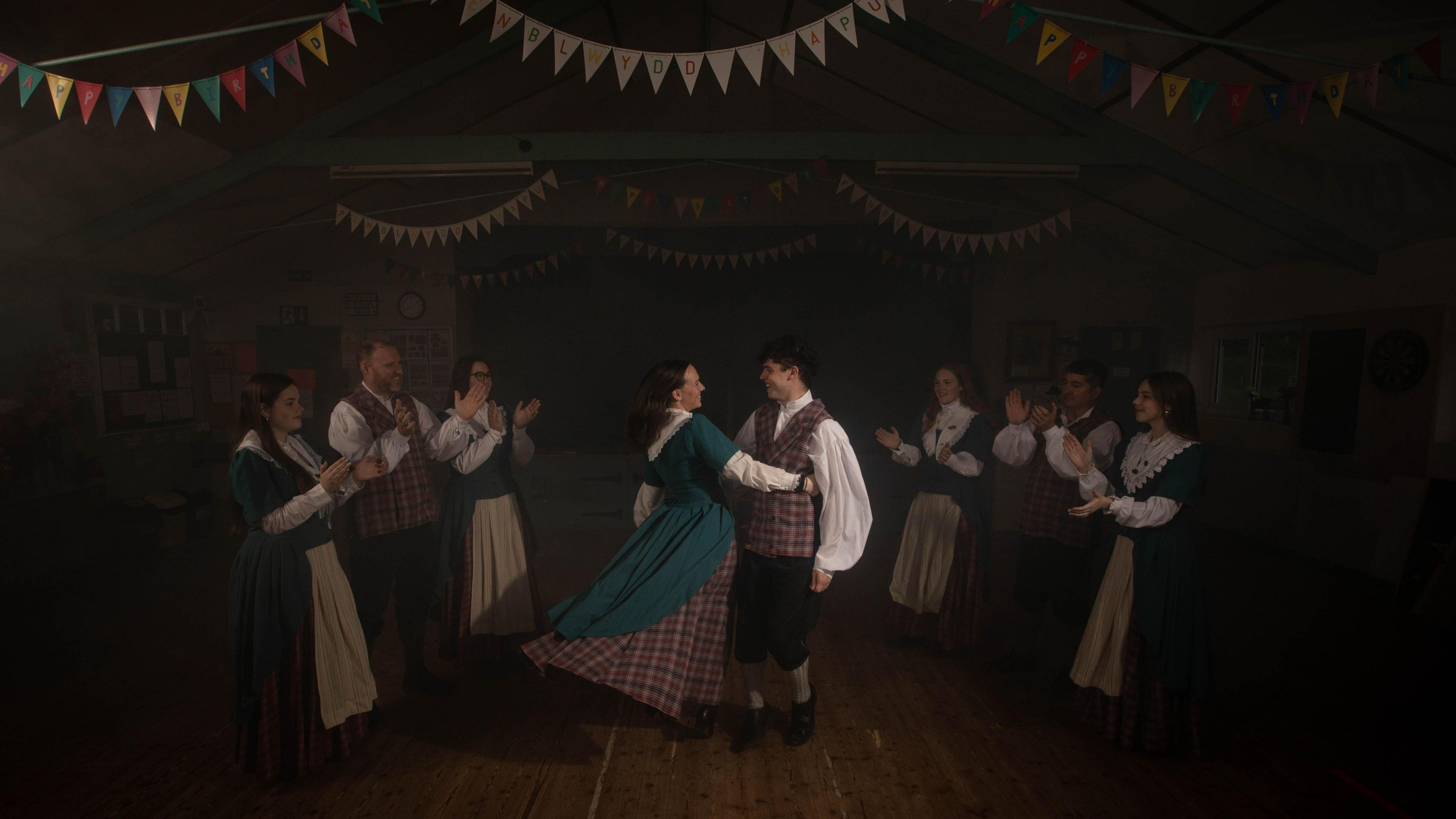 A dark photo. There is a couple in the middle in traditional Welsh attire dancing, the woman holds on to the mans waist. She has a tartan underskirt, with a blue dress on the top, and you can see her lower arms covered in a white sleeved undershirt. The man has a white shirt on and a tartan vest. He has black trousers and khaki socks rolled up with black shoes. There are three people to the left of them clapping, who have the same attire on - one man and two women. On the right there are two women and a man clapping. There are triangular banners strung above the ceiling at the top of the image.