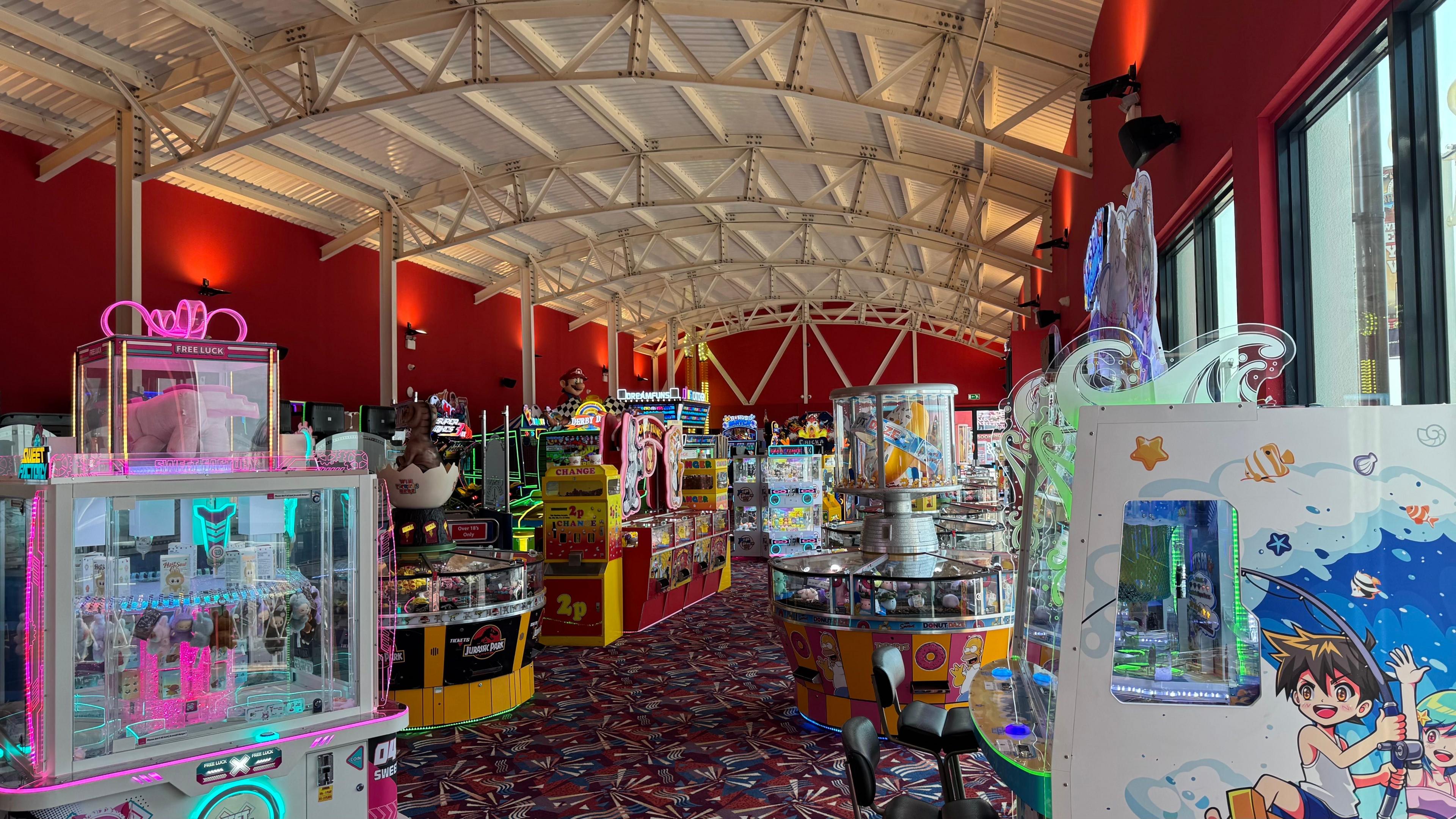 The inside of a small building painted red which has an arched white roof. Multiple bright arcade games are dotted around the room.