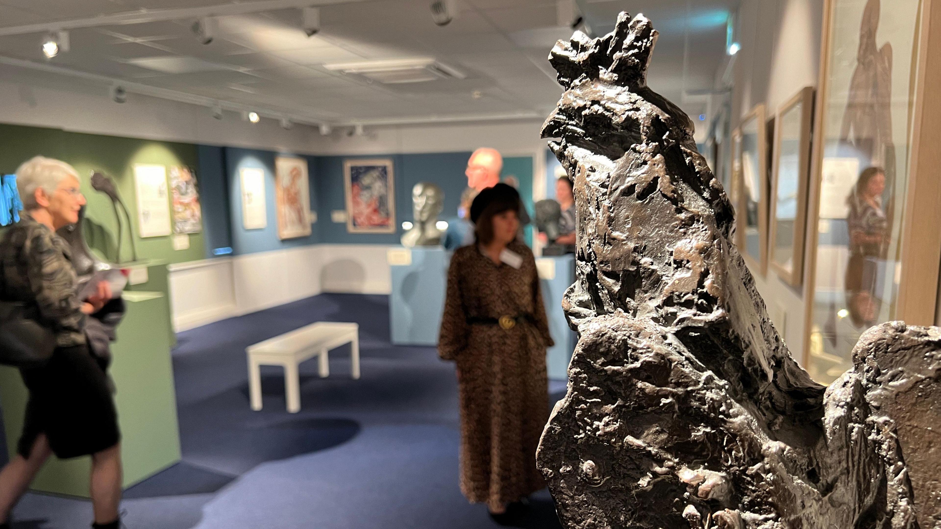 A general view of the interior of the new museum space. A large rock-like formation is shown in the foreground in focus, with out of focus visitors seen looking at the works on display in the background. 