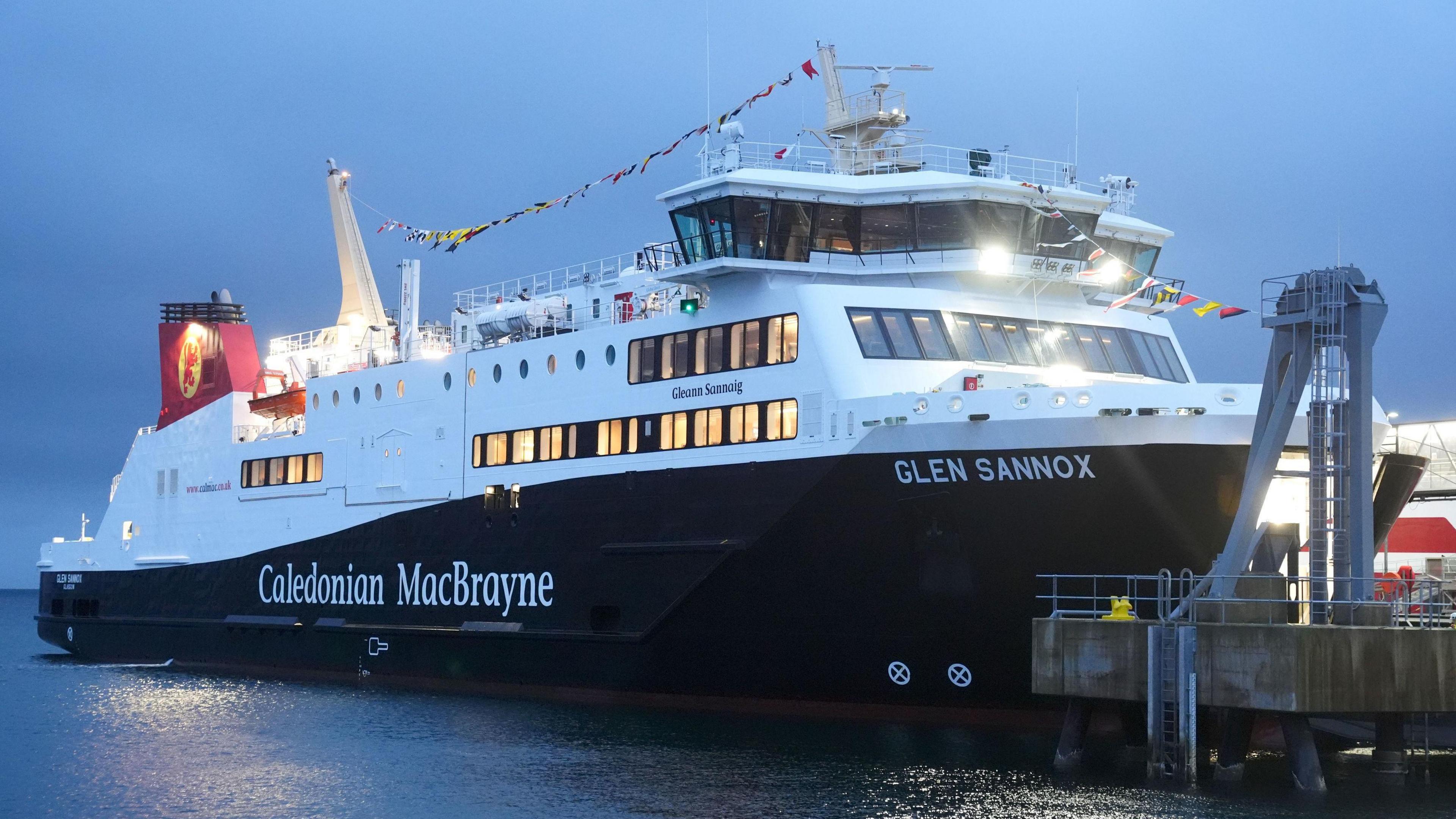 A general view of the Glen Sannox CalMac ferry. The boat's hull is dark coloured with the name of the company and the name of the ship in white writing. The top of the boat is white with a small red section. There are a number of windows on the side with lights on and a row of flags hanging above the passeger deck. It is sitting on calm, dark water on a dark, grey day.