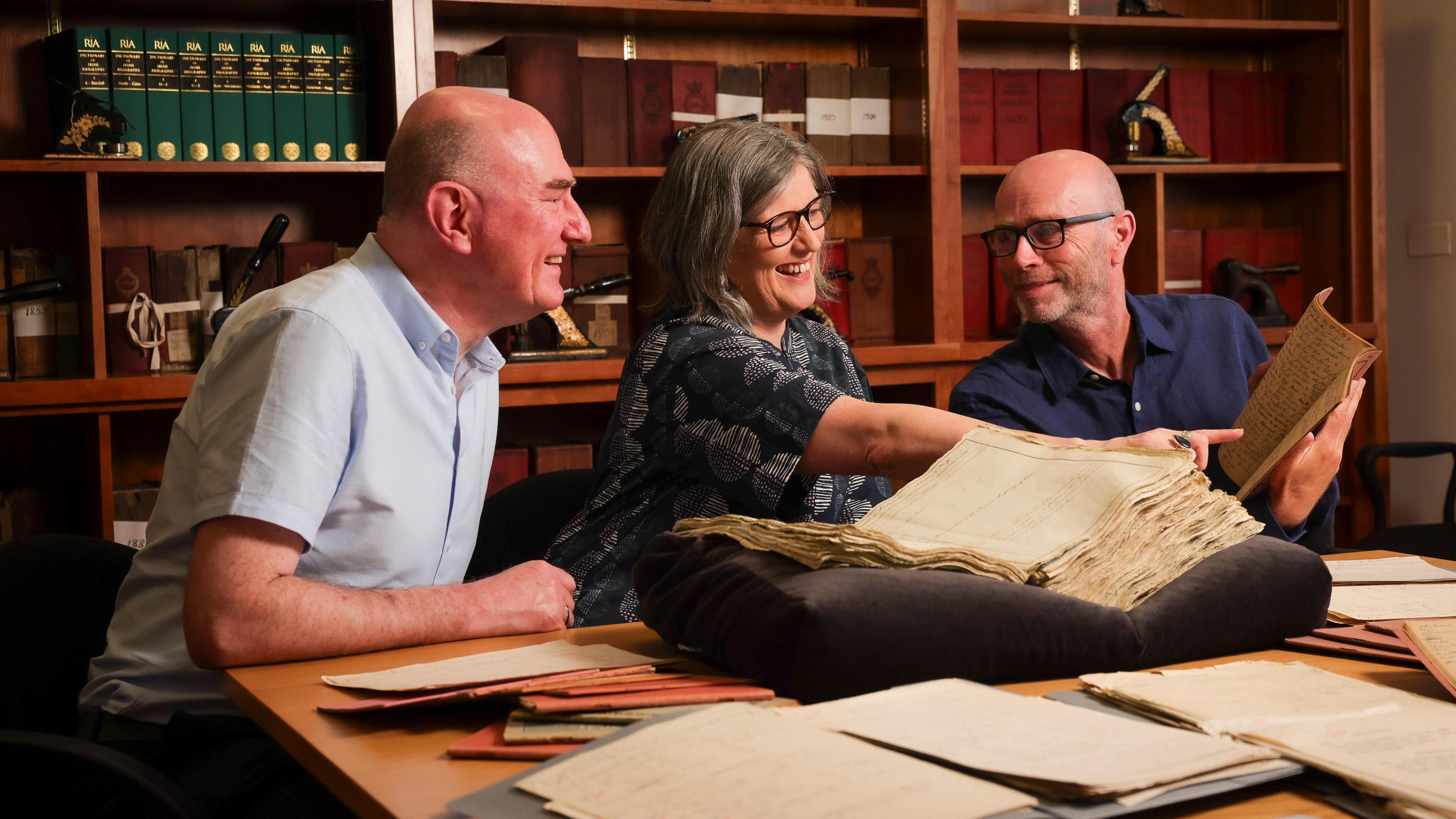 Dr Wallace, left, is looking towards the right of the screen where Dr Gurrin is sitting. He is bald and is wearing a light blue short sleeve shirt. Ms Reid, middle, is reaching toward a book being held by Dr Gurrin, she is wearing a grey top and has short grey hair with black glasses on. Dr Gurrin, right, is holding a book and looking at Ms Reid. He is wearing black framed glasses, a navy shirt and is bald.