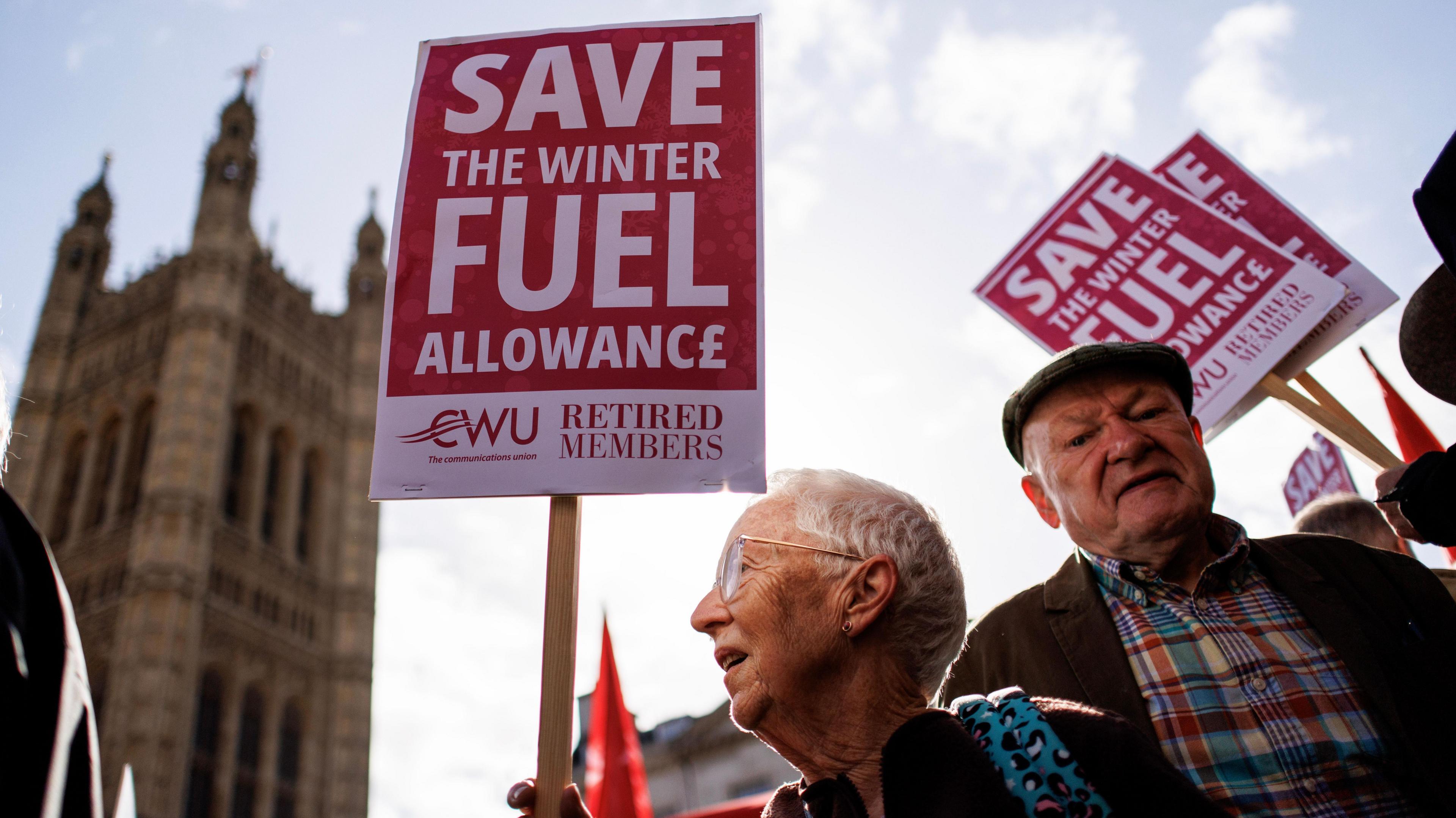 An elderly looking man and woman holding up placards saying 'Save the Winter Fuel Allowance'. The Houses of Parliament can be seen in the background.