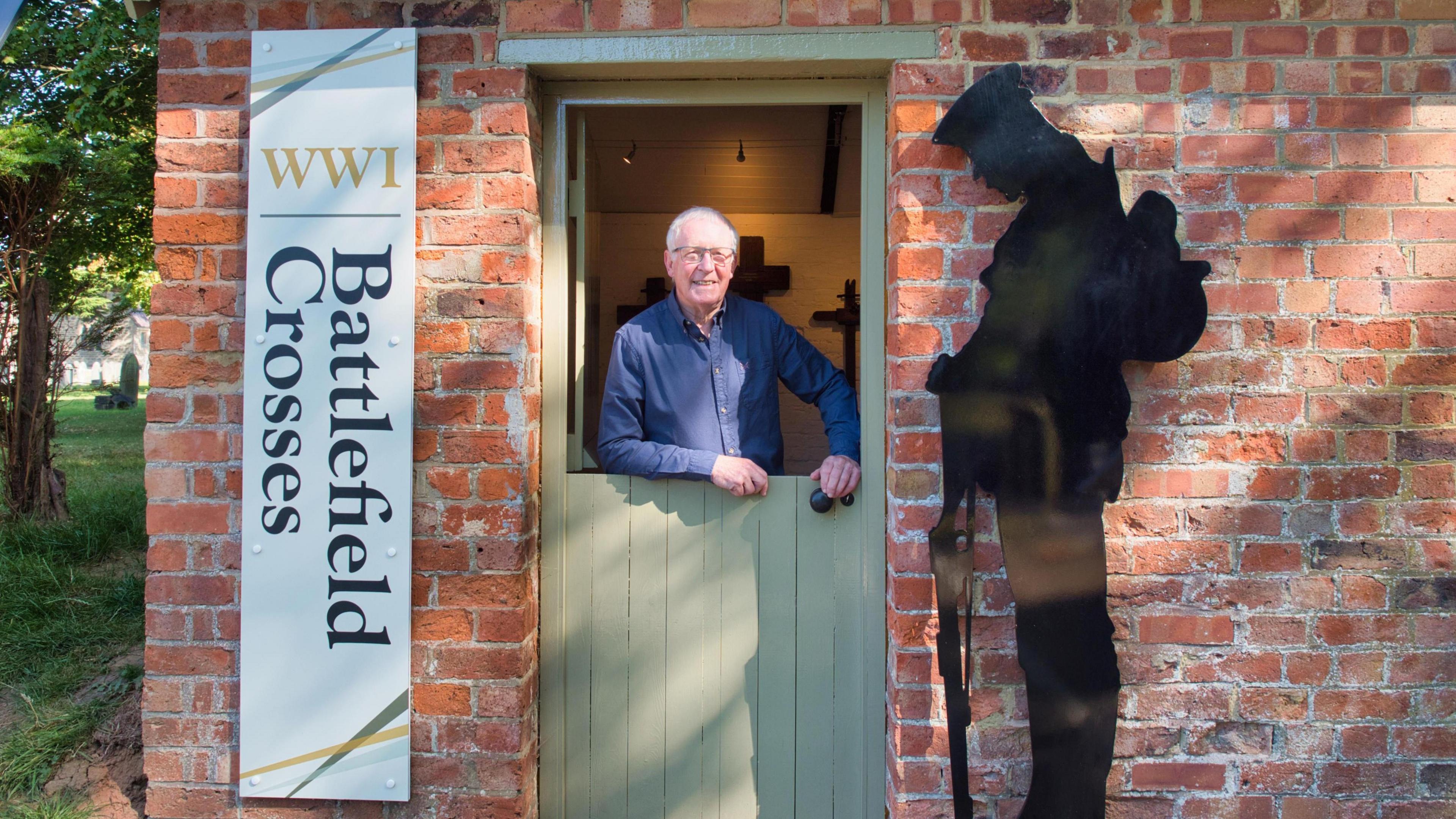 A man with white hair and glasses standing behind a stable door which is closed on the bottom. He is standing in a small brick outhouse building with a vertical sign on the left that says 'World War One battlefield crosses', with a silhouette of a soldier on the right of the door.