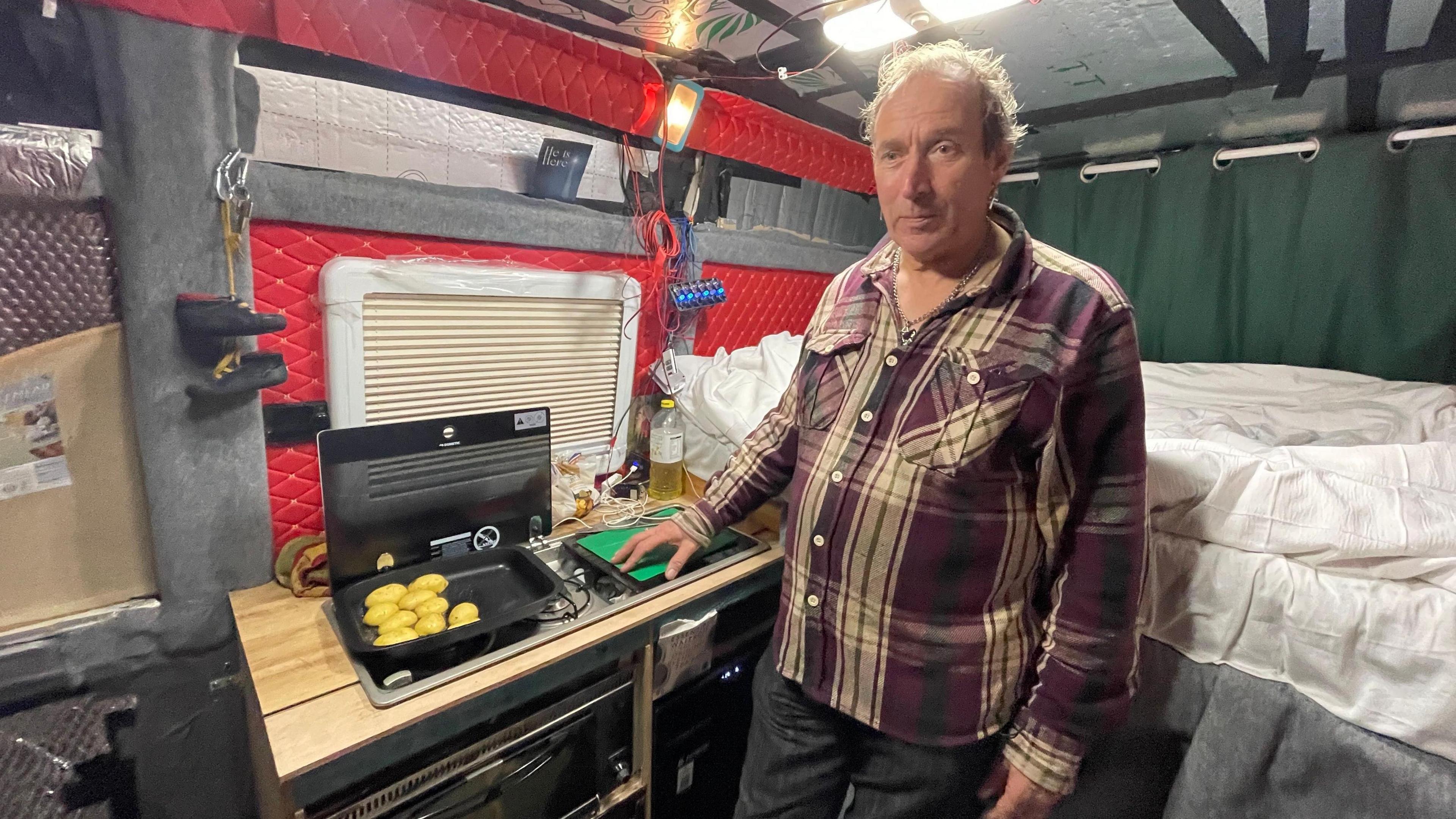 Dave Knight is standing inside his caravan cooking potatoes. A bed is right behind him and he is wearing a checked shirt.