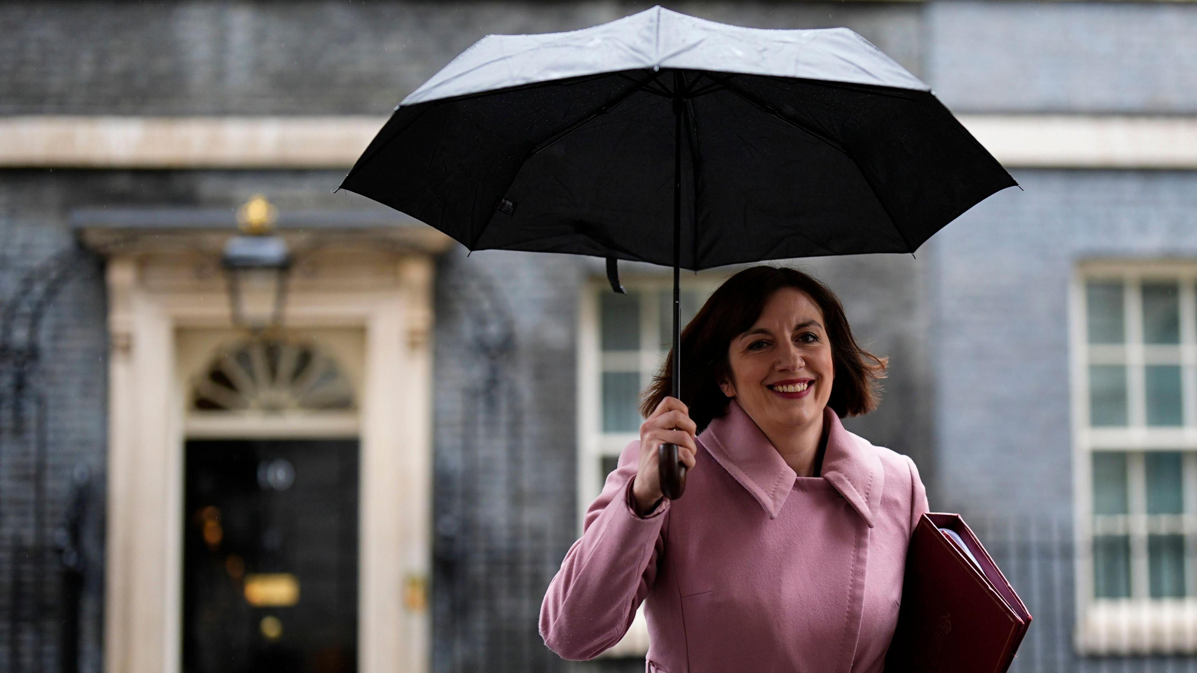 Bridget Philipson walking out of downing street smiling and holding an umbrella and a wearing a dusty pink coat.
