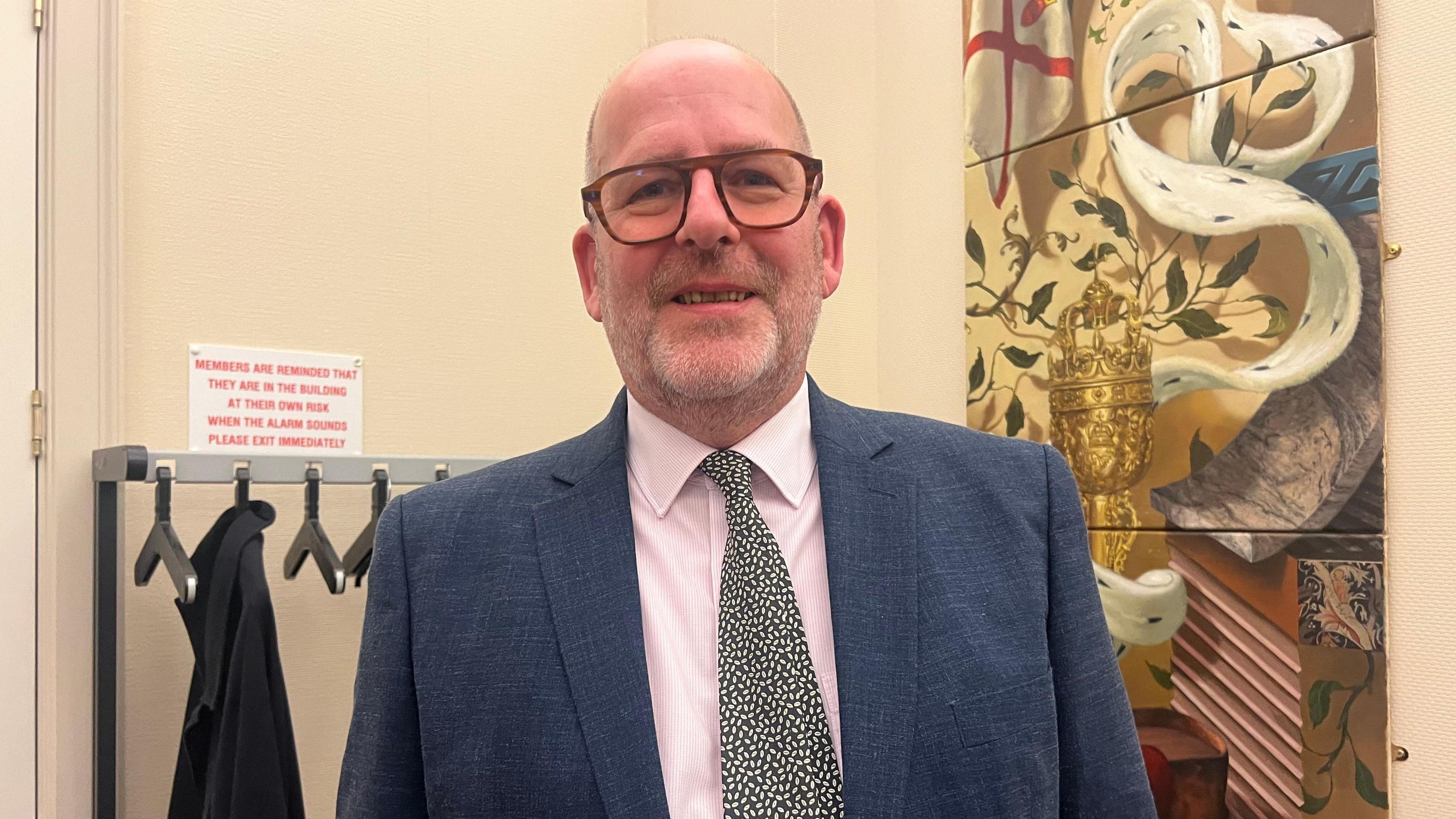 Professor Andrew Le Sueur is smiling at the camera. To his left is a painting showing a chalice and the Jersey flag and behind him is a coat rack. He is wearing glasses and a pink shirt, patterned tie and navy jacket. 