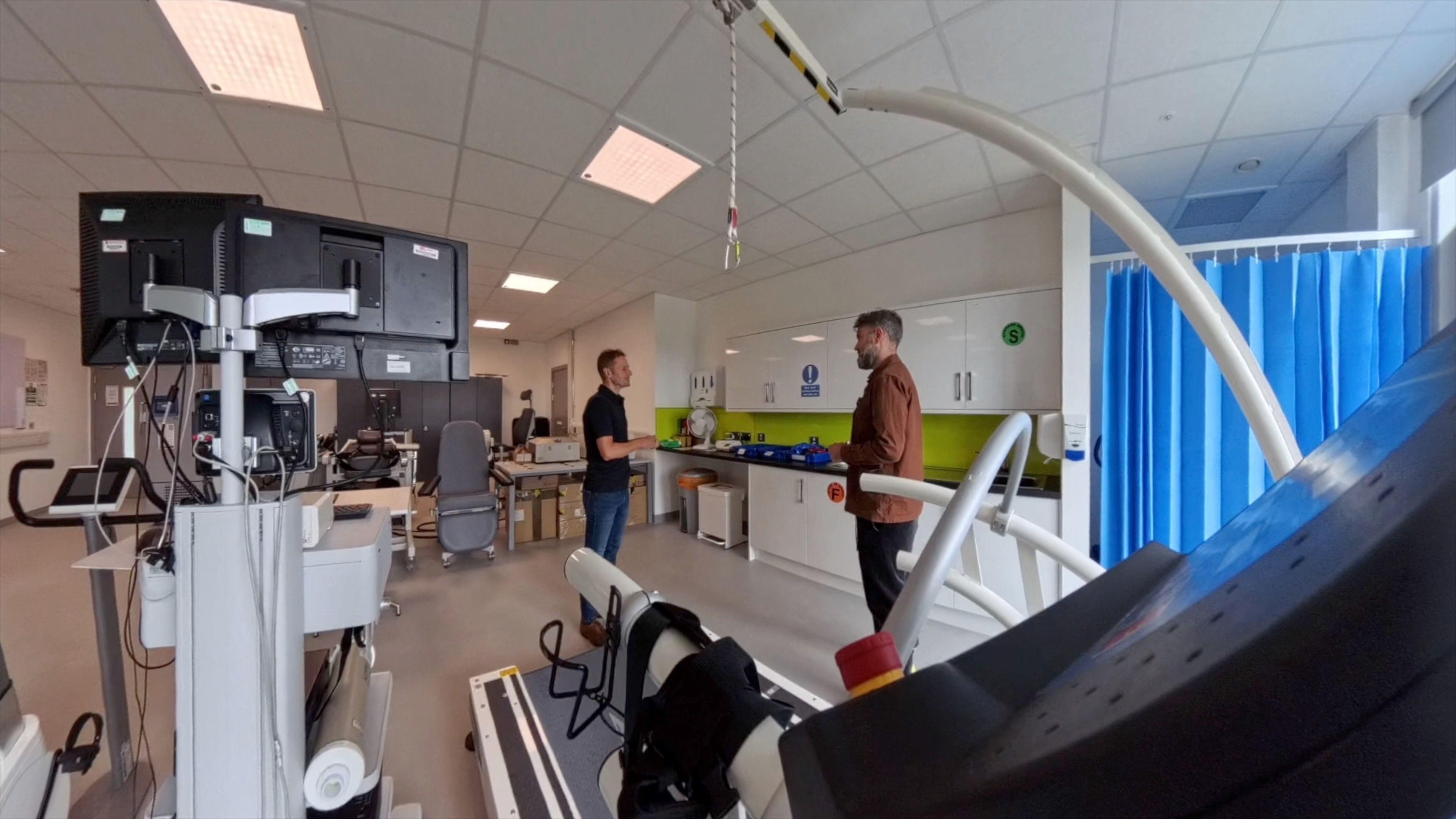 Rob Copeland from the Advanced Wellbeing Research Centre talks to BBC Yorkshire's James Vincent in one of their labs. Rob is wearing a dark polo shirt - James is wearing a Brown shirt. they are standing behind a treadmill used for analysis of athlete performance.