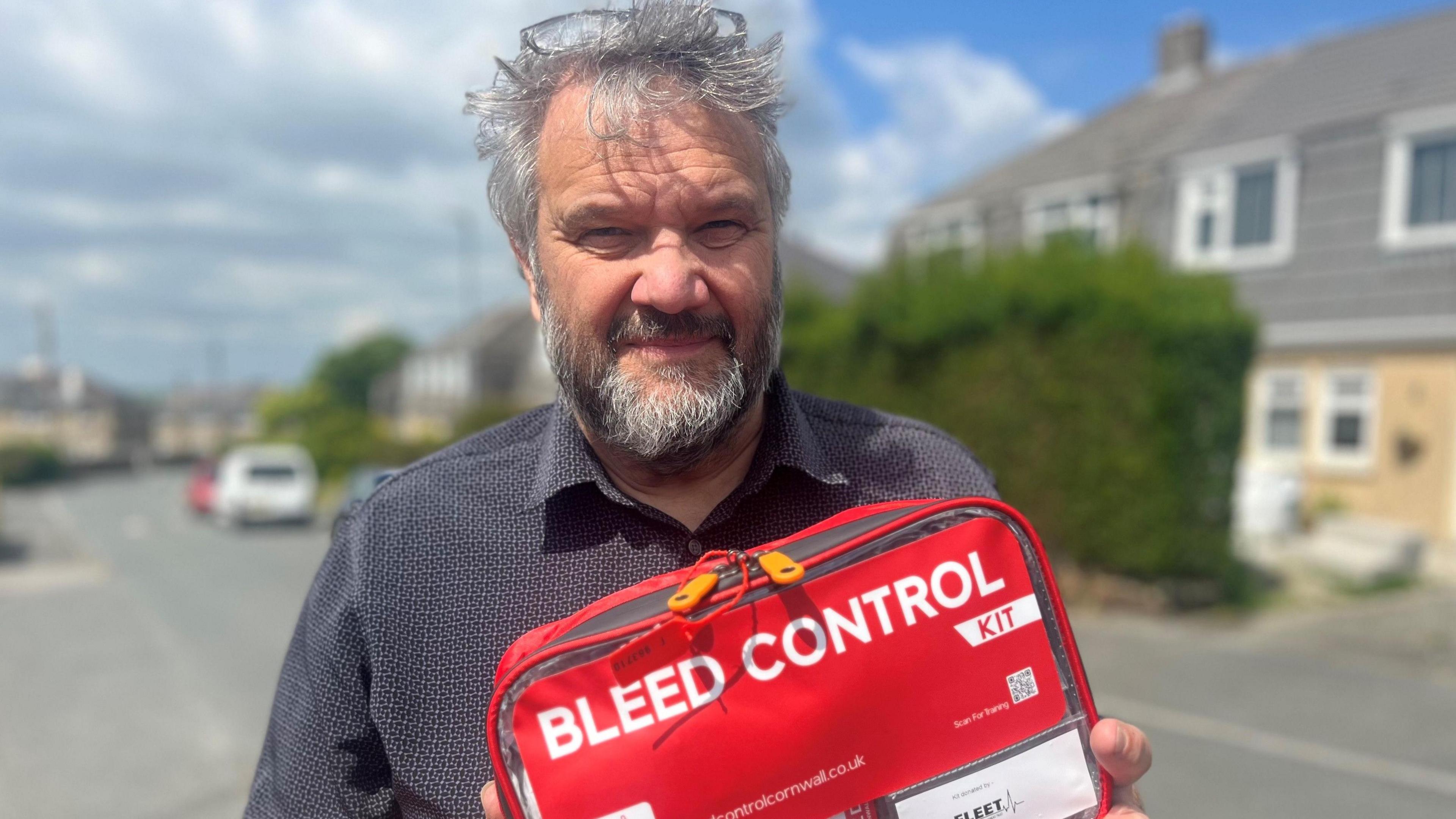A man with grey hair and a beard looking at the camera, he is wearing a dark grey shirt and has glasses pushed back on his head. There is a street with houses in the background and he is holding a red case which says bleed control kit in white lettering