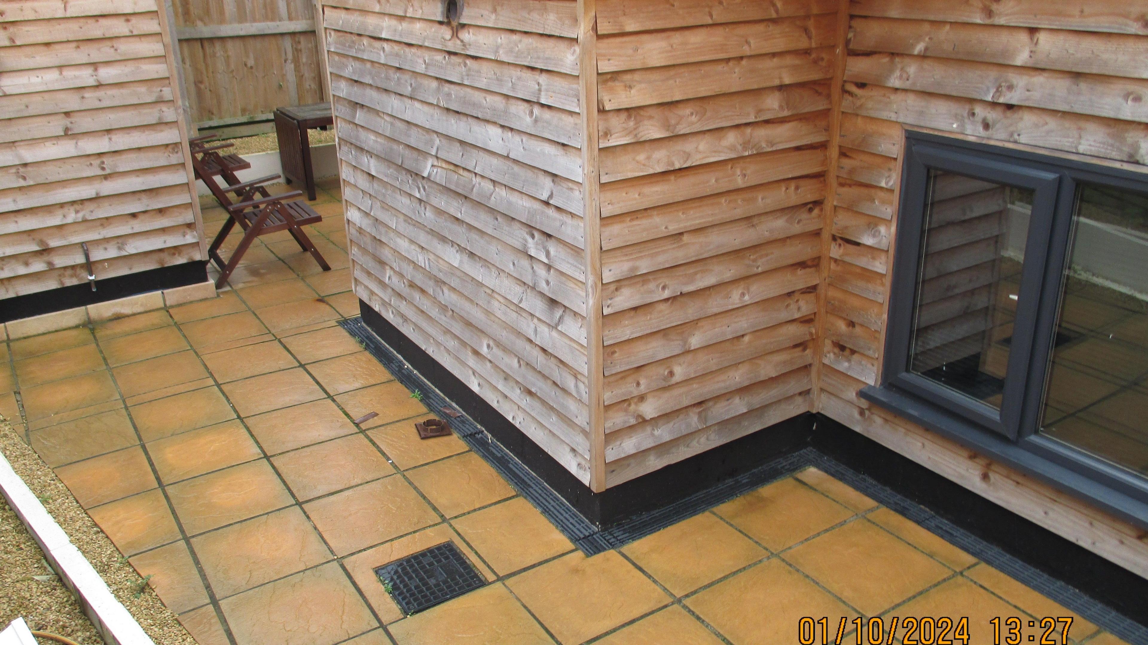 The side of one timber building and the corner of another with a yellow patio area in between. A window can be seen on one of the buildings. A pair of patio chars and the edge of a table can be seen between the buildings.