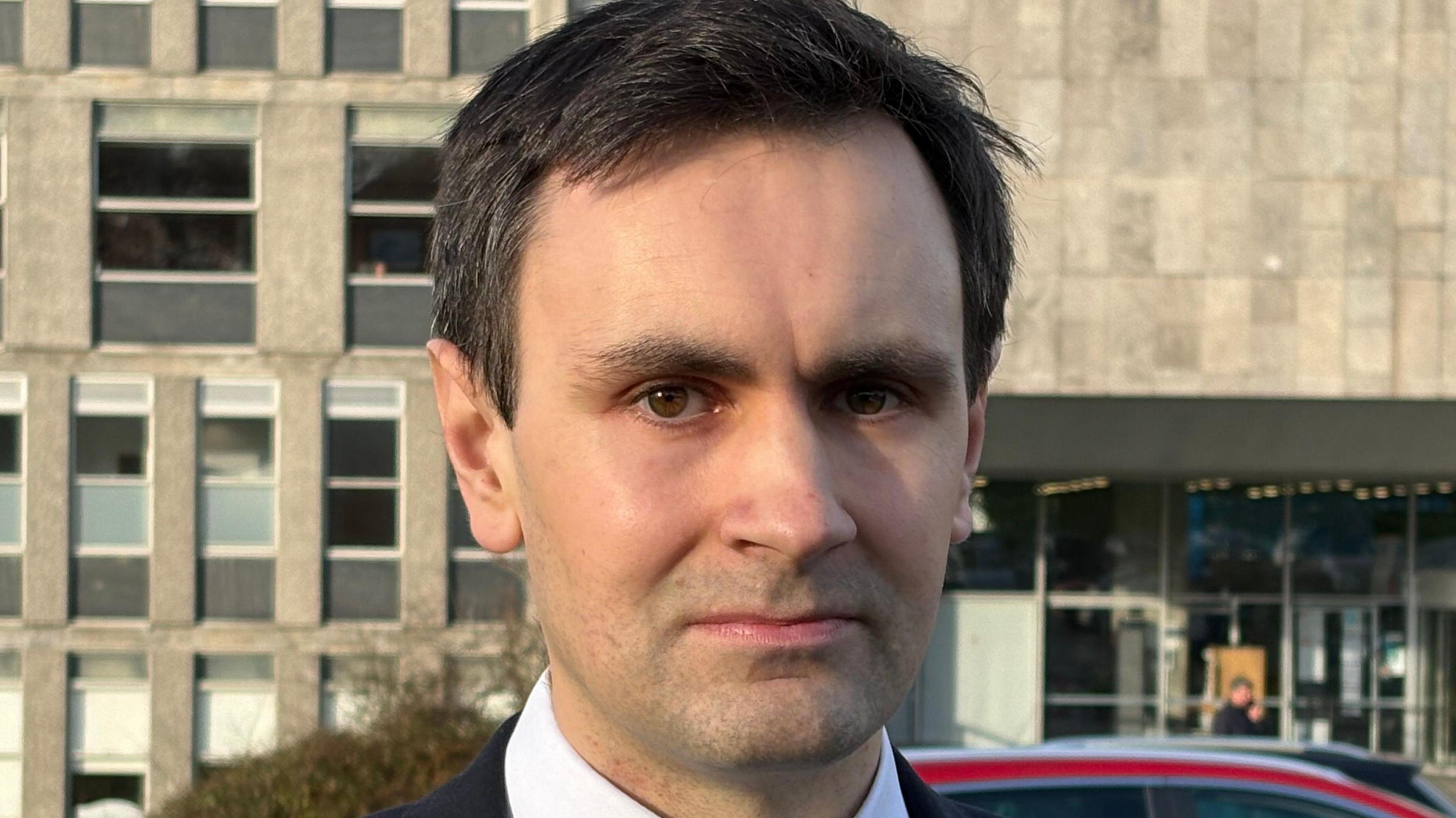 Man with dark hair in white shirt standing in front of Cornwall Council's HQ