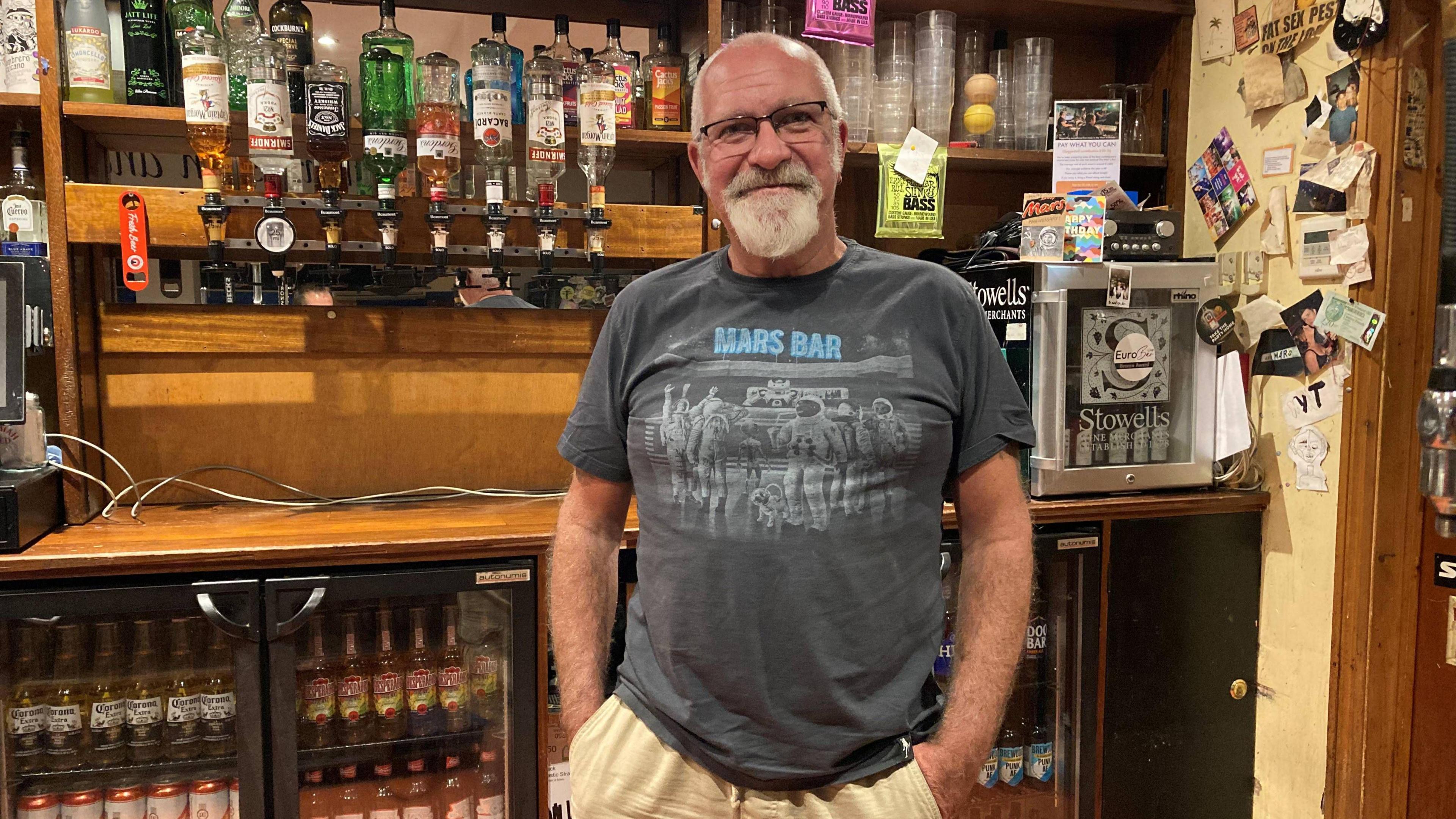 A man wearing a t-shirt that says 'Mars Bar', stood behind a series of drink cabinets full of bottles of beer.