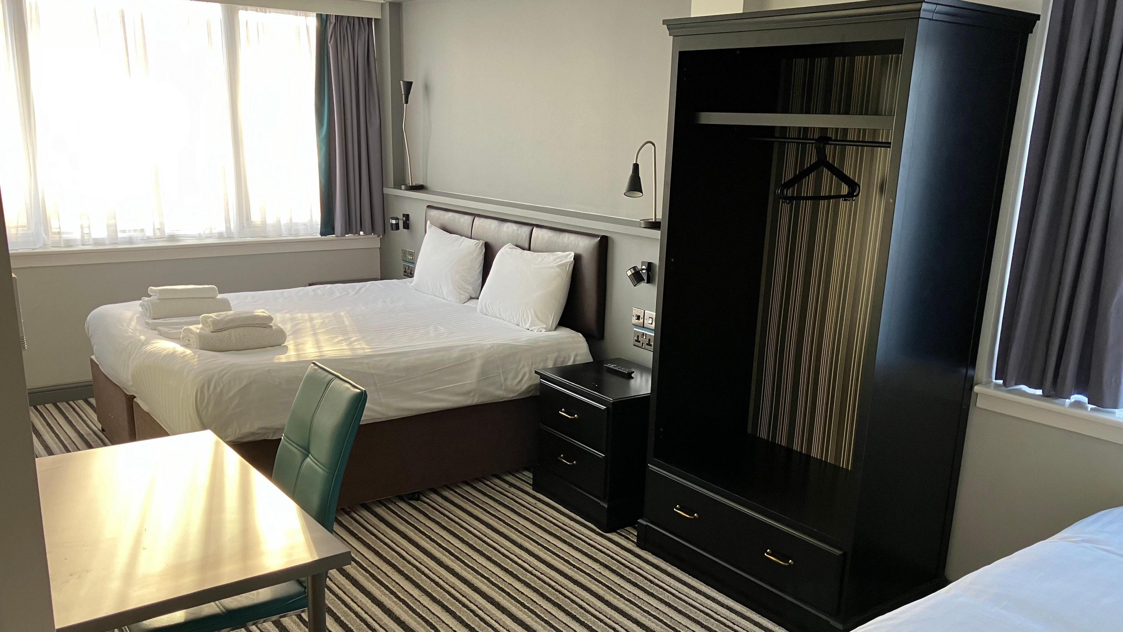 An empty hotel room showing a double bed with white sheets and a wardrobe. Part of a single bed can be seen, as can a table. There is a striped carpet and curtains. 