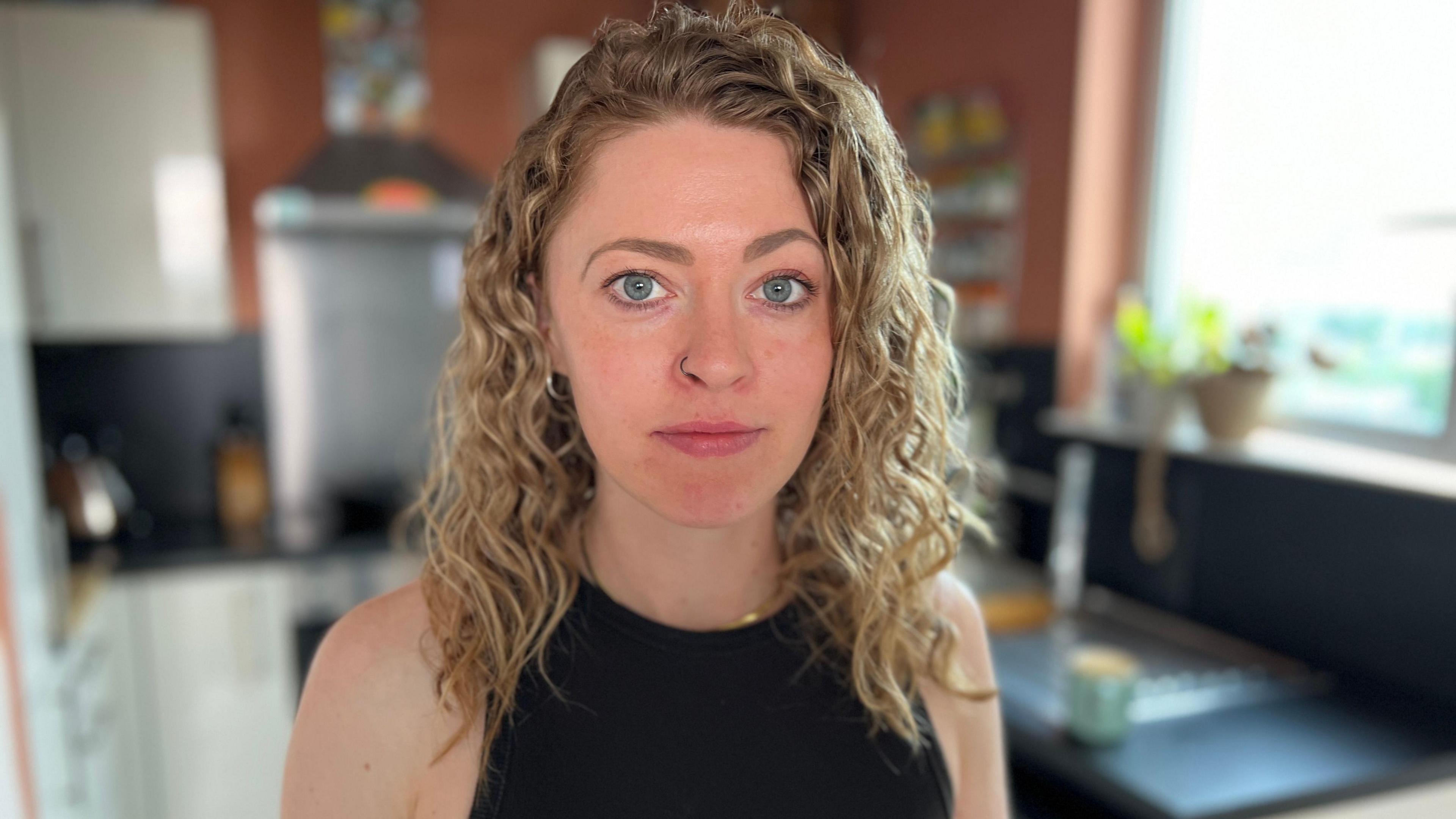 Rosanna Heasman looks directly at the camera standing in a kitchen, she has curly blonde hair and is wearing a black top