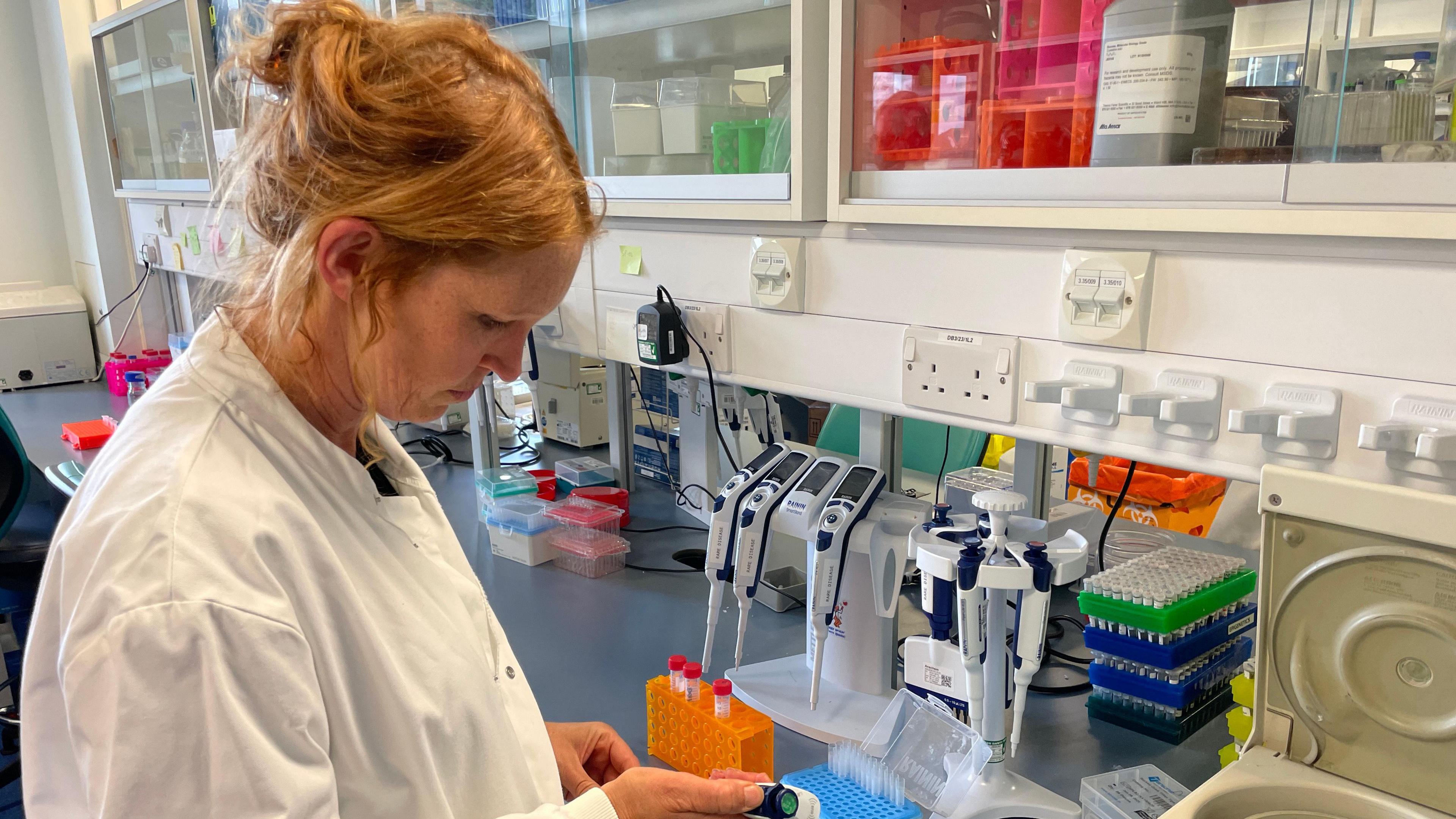 Woman with light auburn hair in a white lab coat holding test tubes in a laboratory, she is looking down at them side profile