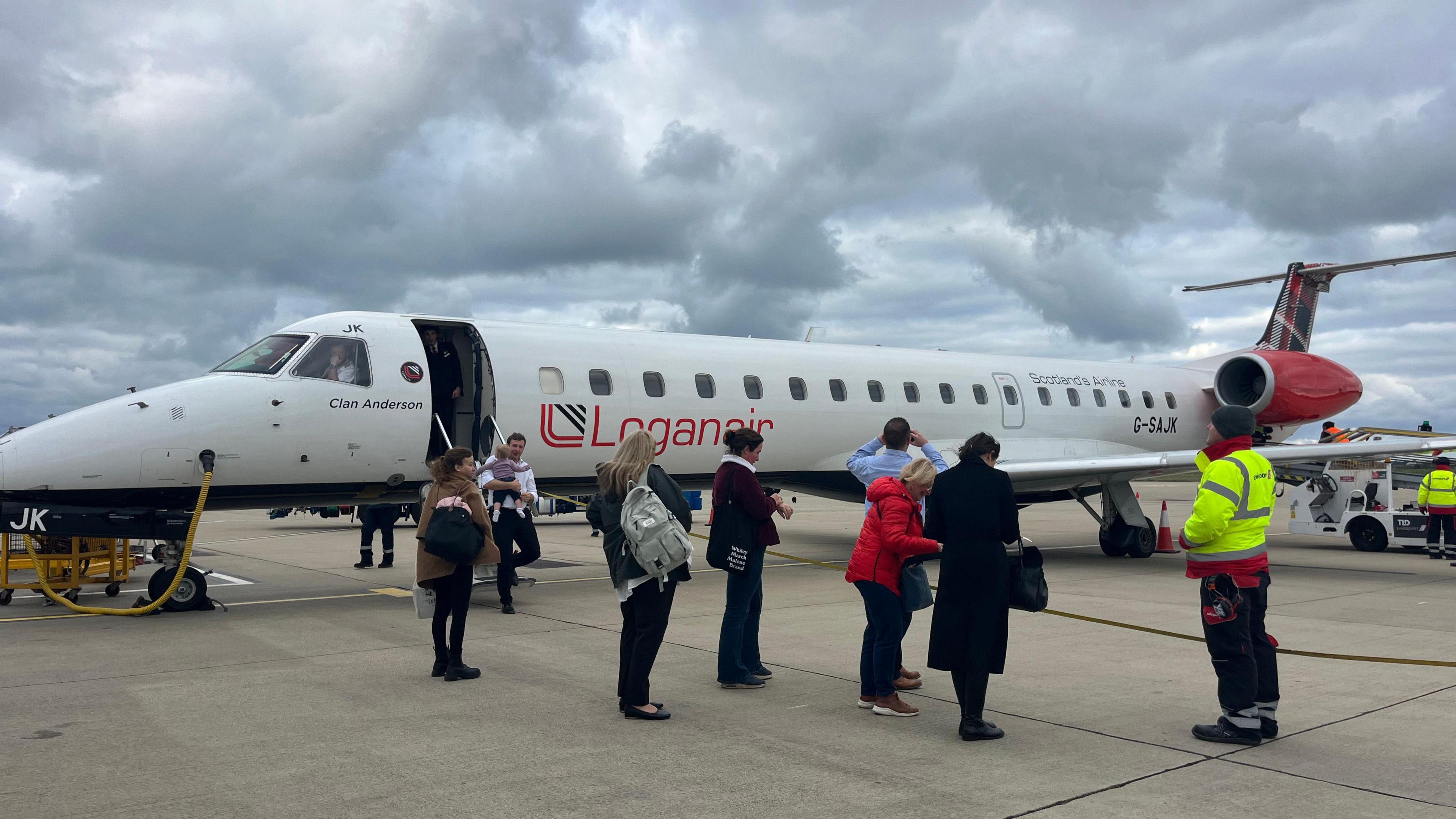 The Loganair-branded plane is on the tarmac. There are seven passengers on the ground and one man in a high viz air crew jacket. It is a grey day.