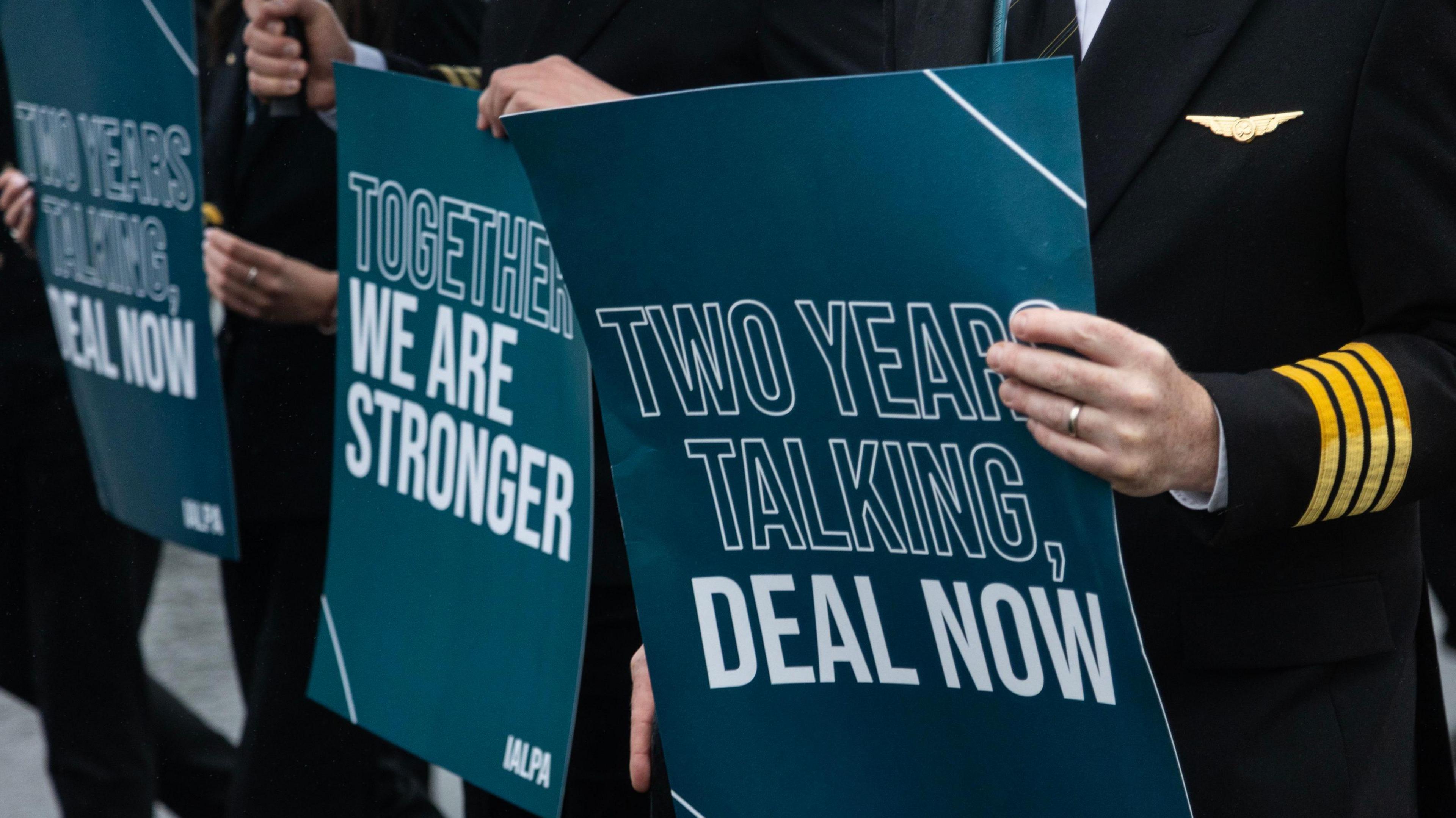 Pilots on strike in June, picture is close up of them holding blue signs with white writing which read 'two years talking, deal now' and 'together we are stronger'