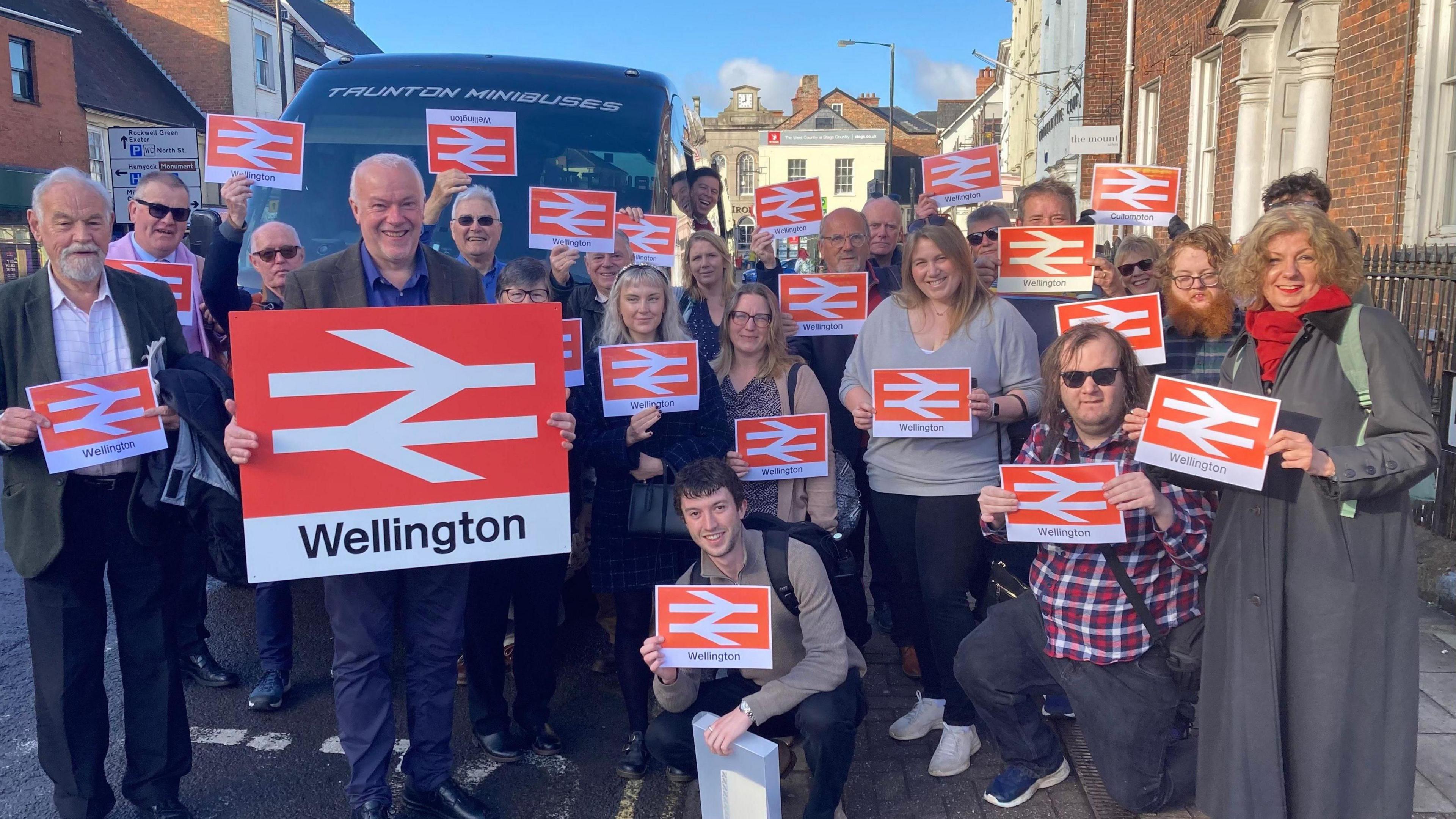 A group of people, standing next to a minibus, holding railway station signs saying "Wellington"