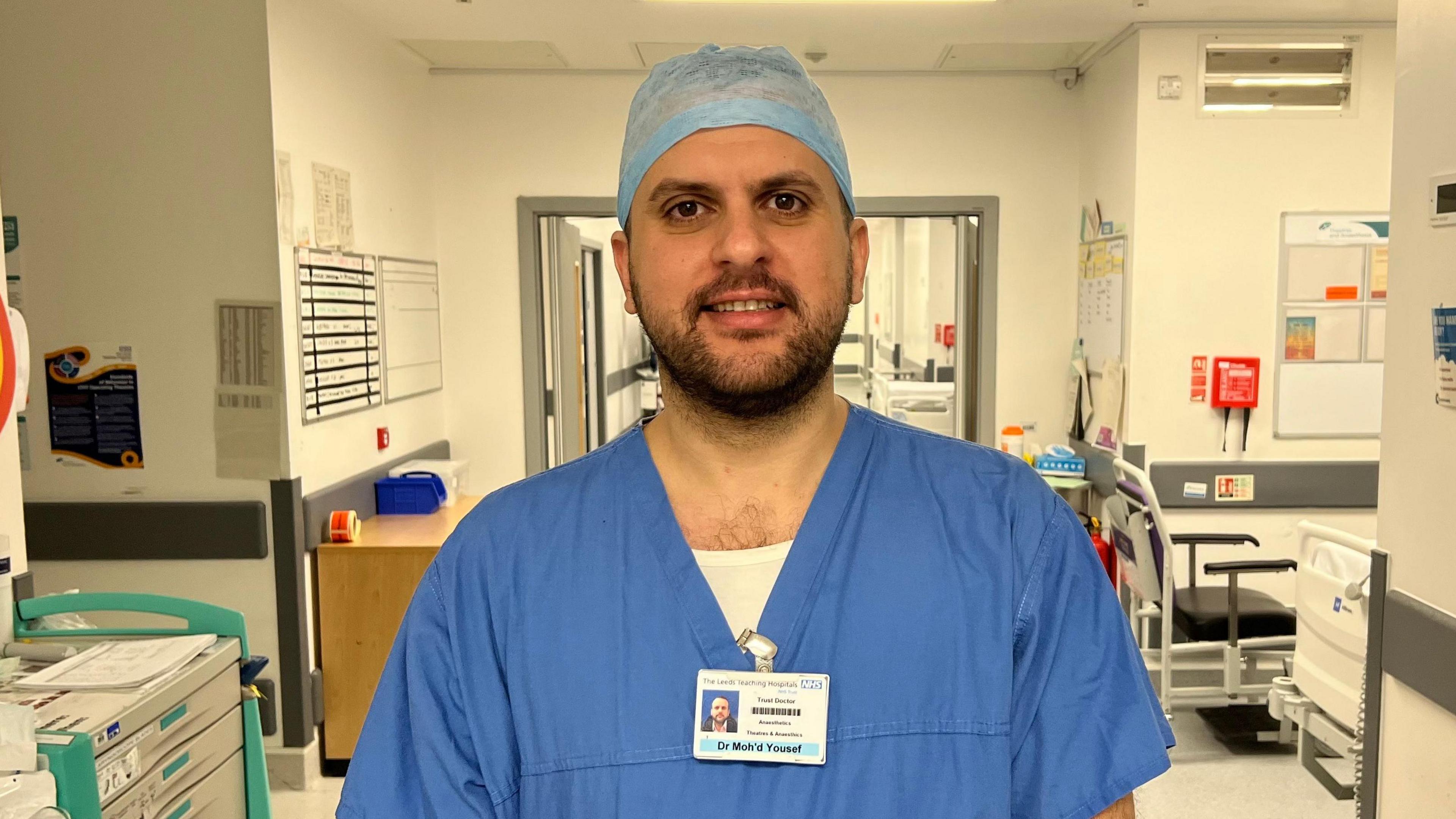 A man wearing blue surgical scrubs is standing in the middle of a hospital corridor.  He is in his thirties and has joined the NHS from Jordan.  He has a short black beard and a name badge attached to his top.