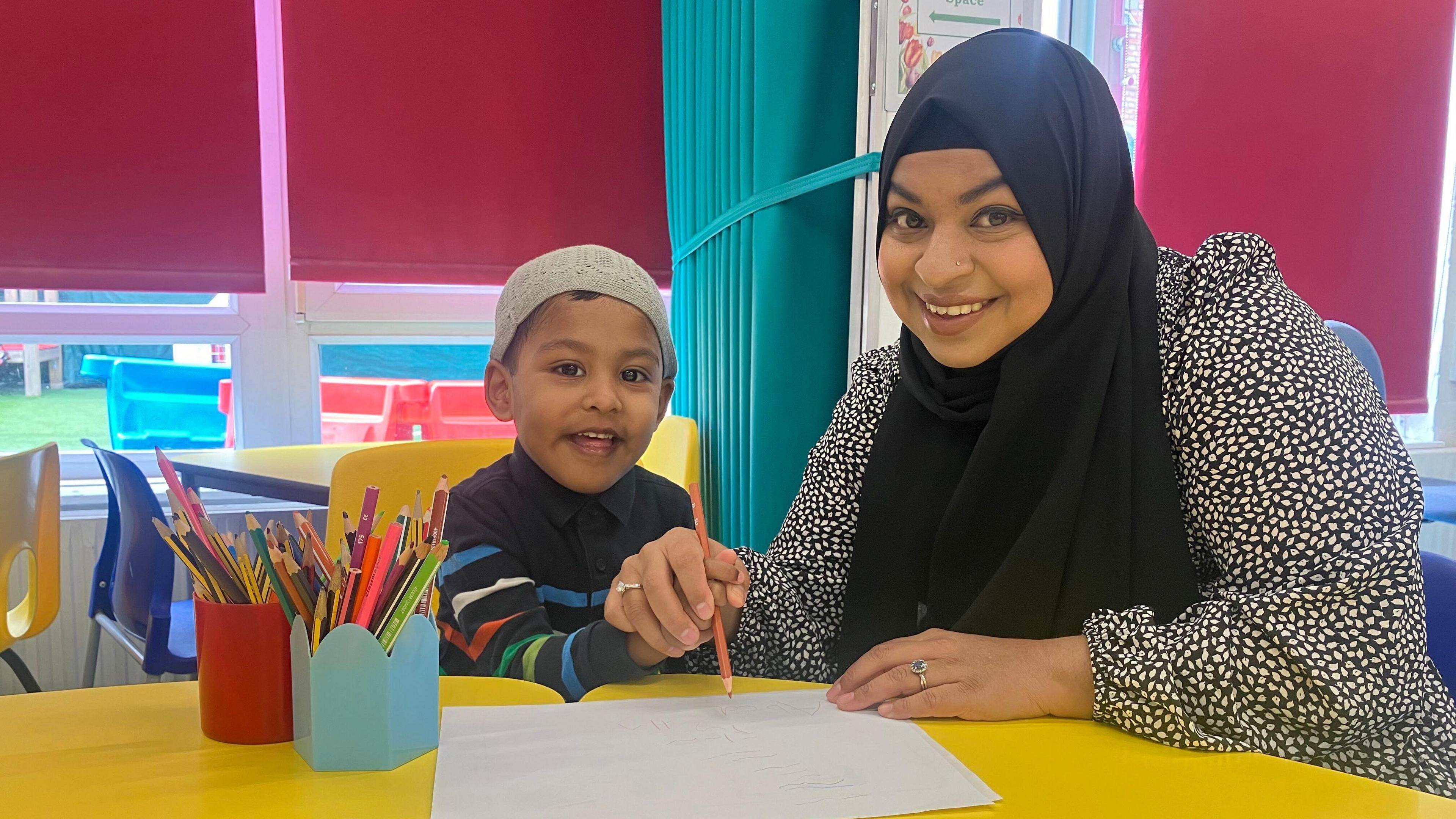 A boy in a grey hat and a stripy navy jumper sits beside a lady in a black and white dress and a black hijab. They are both holding an orange pencil and writing. They are in a classroom sitting at a yellow table with yellow seats and red blinds behind them