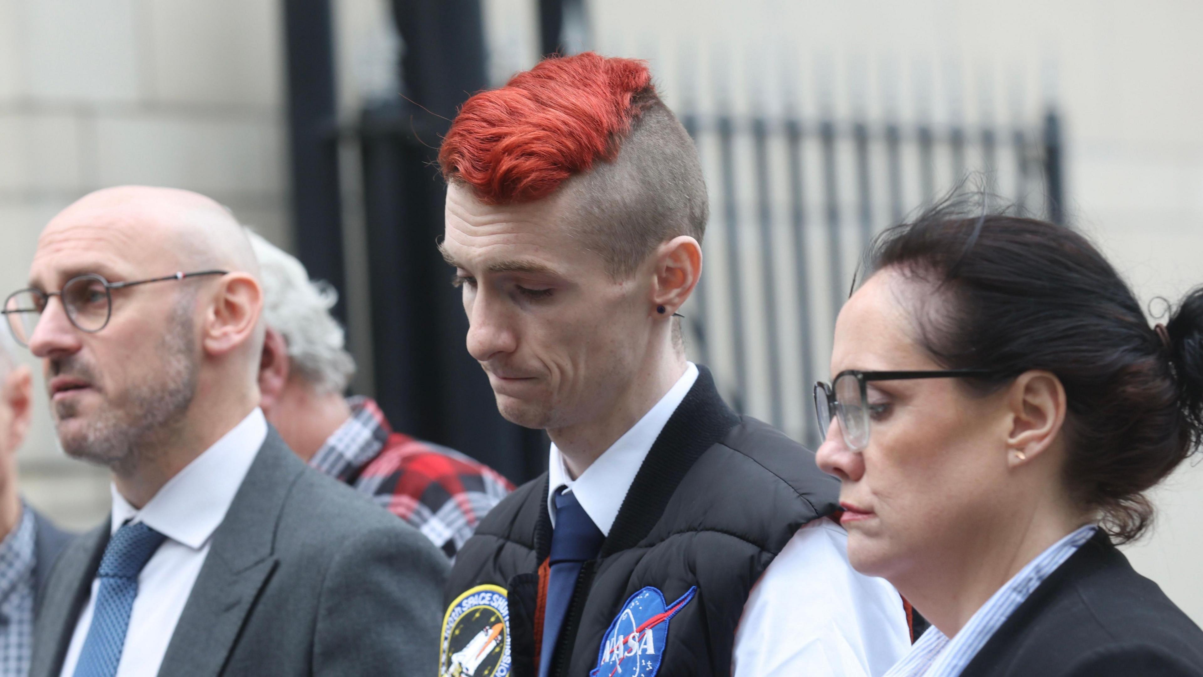 Mr Doyle is looking down to the ground with a sad expression. He has a his hair died red, is wearing a white shirt, black tie and black body warmer