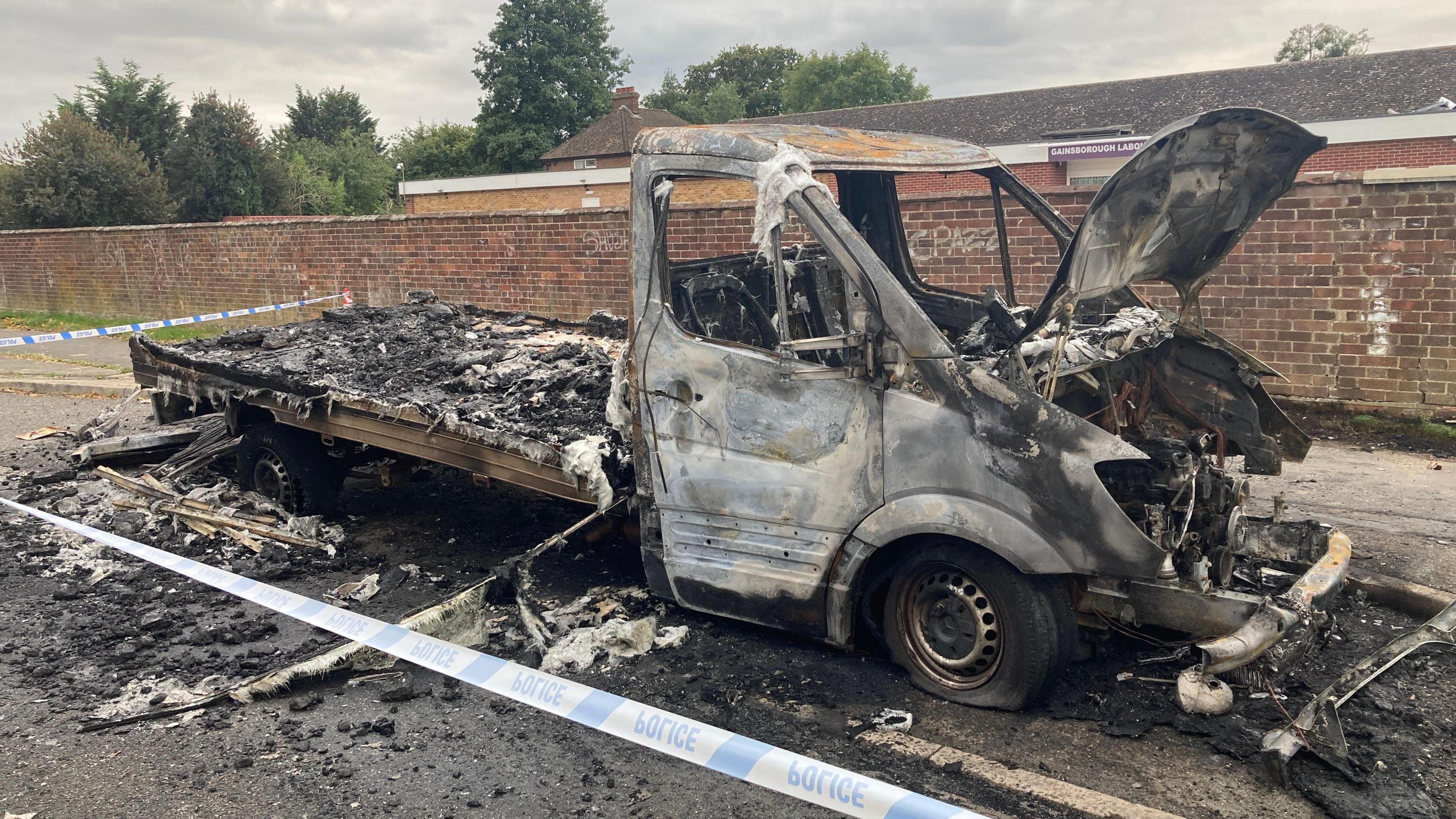 A flat bed van destroyed in a fire within a car park. It is surrounded by police cordon tape. Burnt rubble and metal surrounds the vehicle on the ground.
