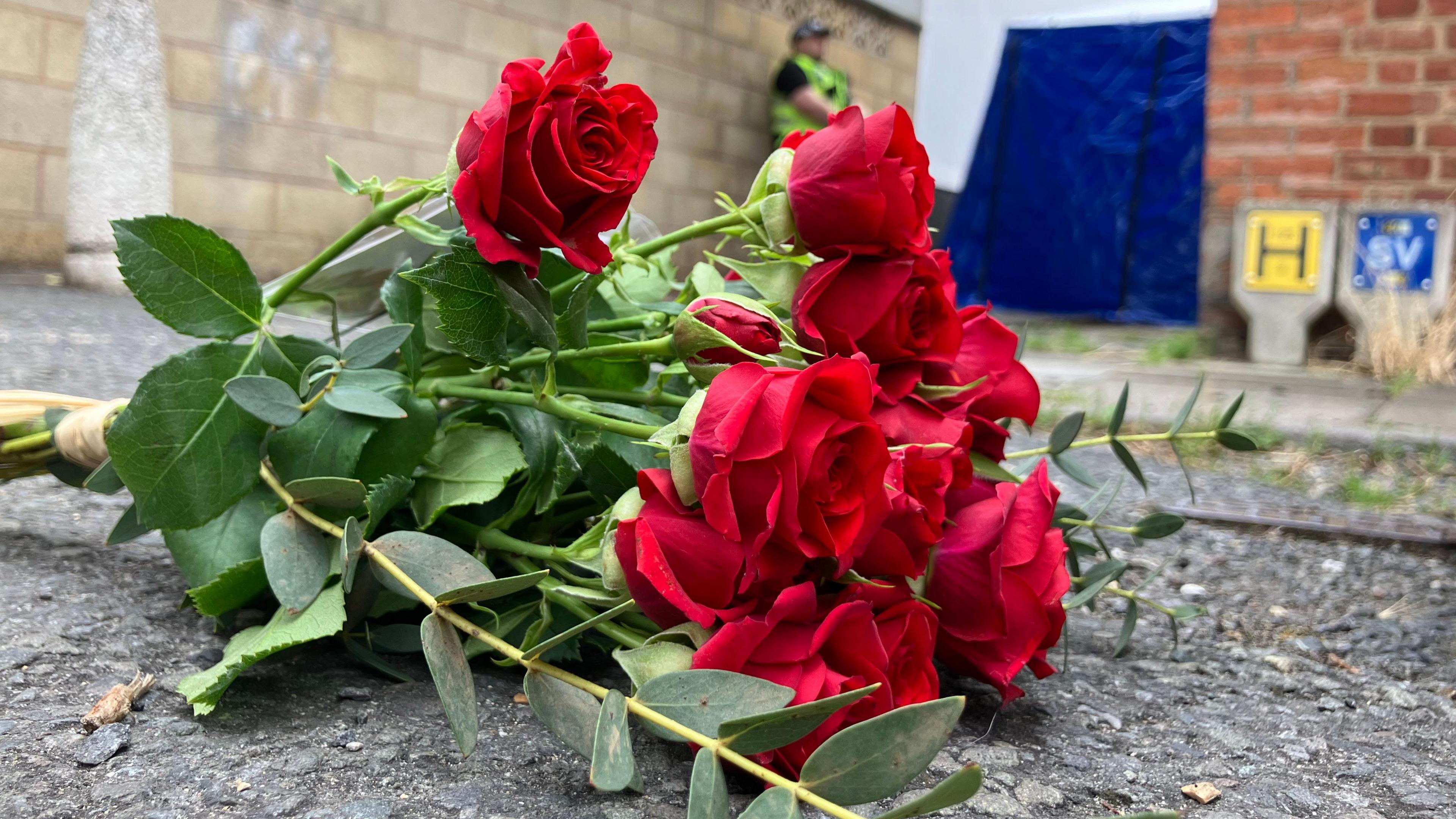 A bunch of roses lie on the concrete floor beside a brick wall. A police officer wearing a high vis jacket can be seen in the background standing next to a blue tent.