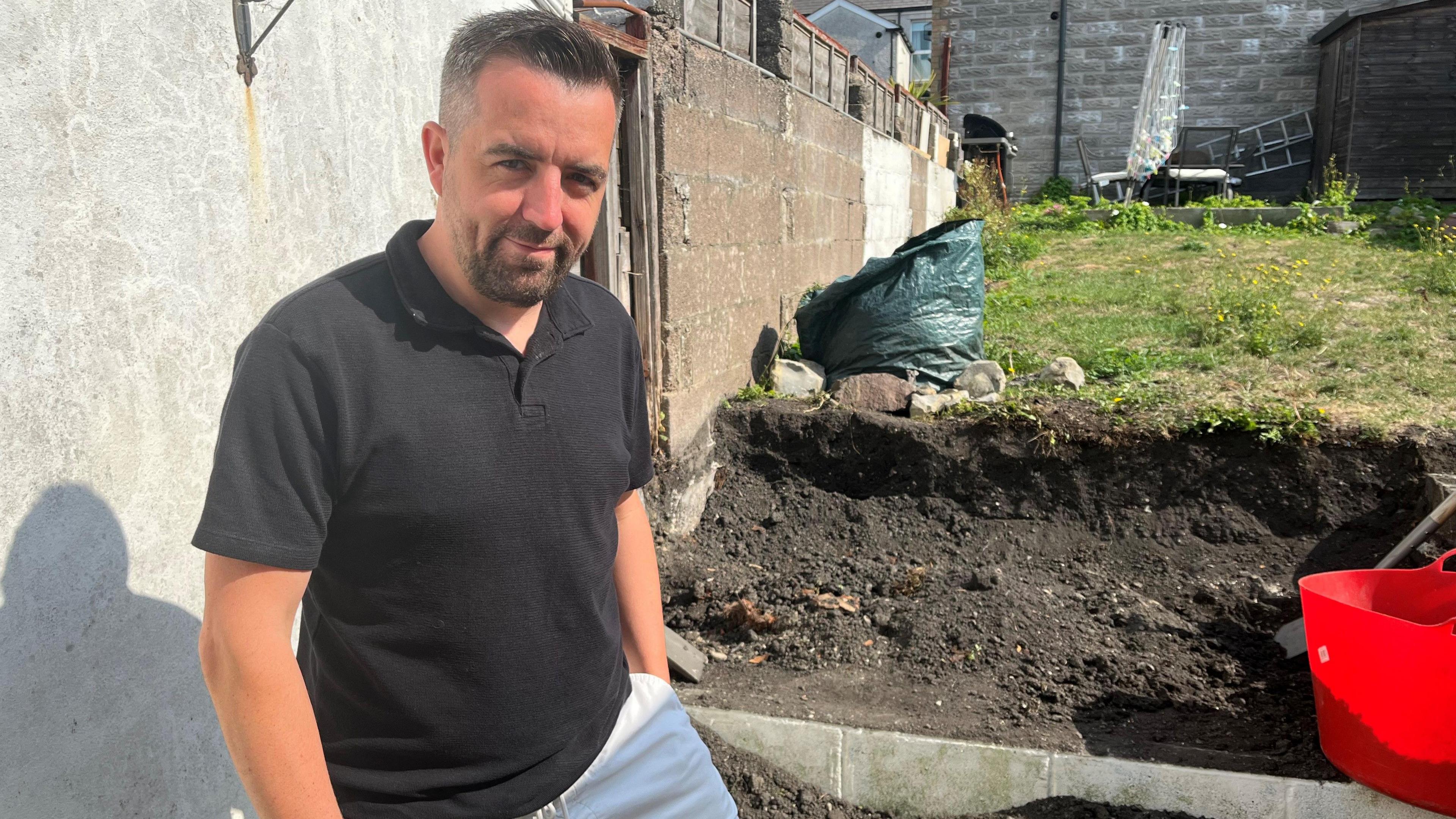 Tom Clarke, who has dark hair with silver speckles and a dark beard, smiles at the camera. He wears a black polo shirt and white shorts and his dug up garden can be seen in the background.