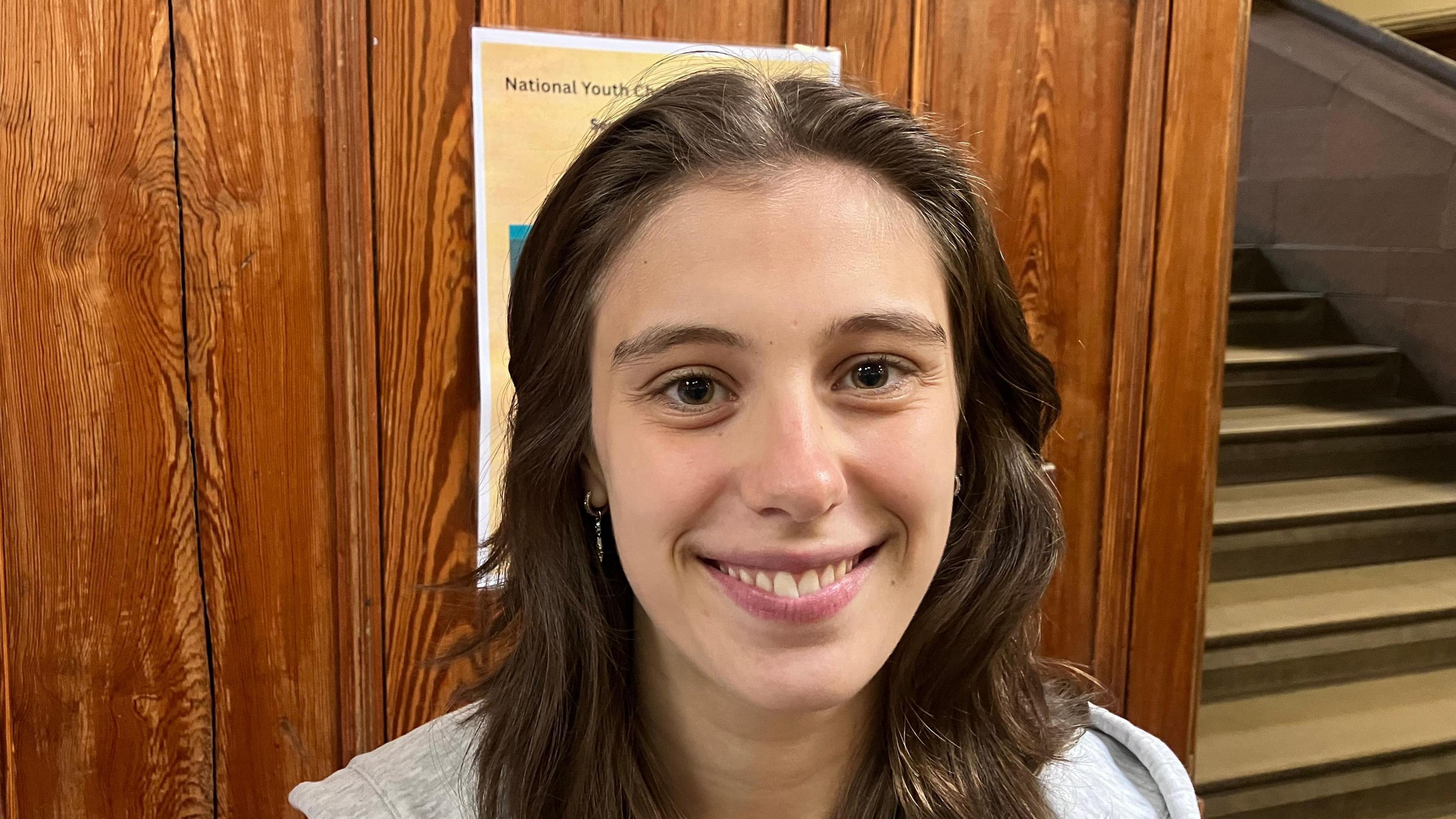 Amy Patton has long brown wavy hair and brown eyes. She is smiling at the camera in front of a brown wooden wall and cream staircase. 
