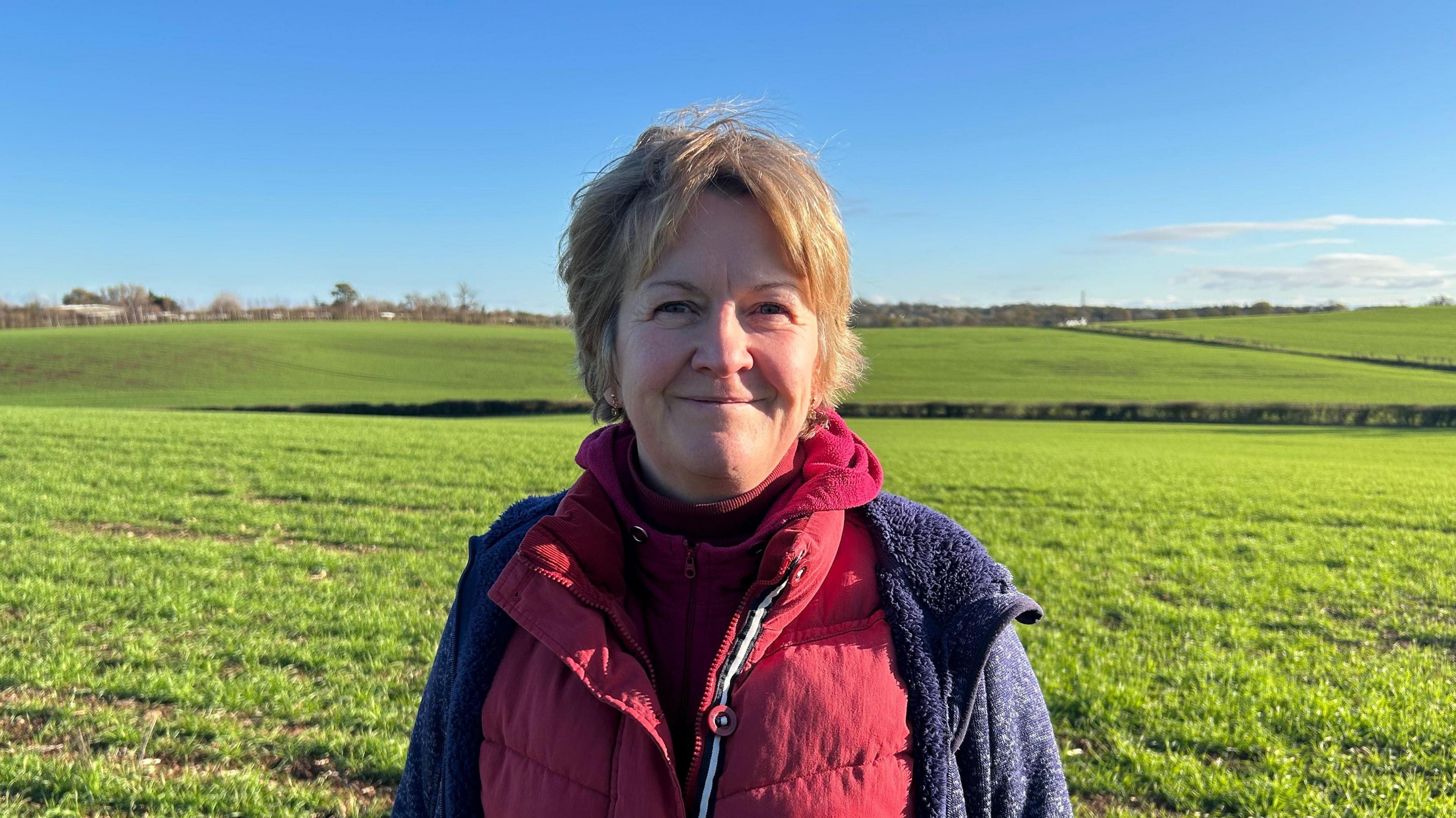 A woman with short light brown hair is wearing a red coat with a blue fleece over the top. Behind her are fields and trees as far as the eye can see. 