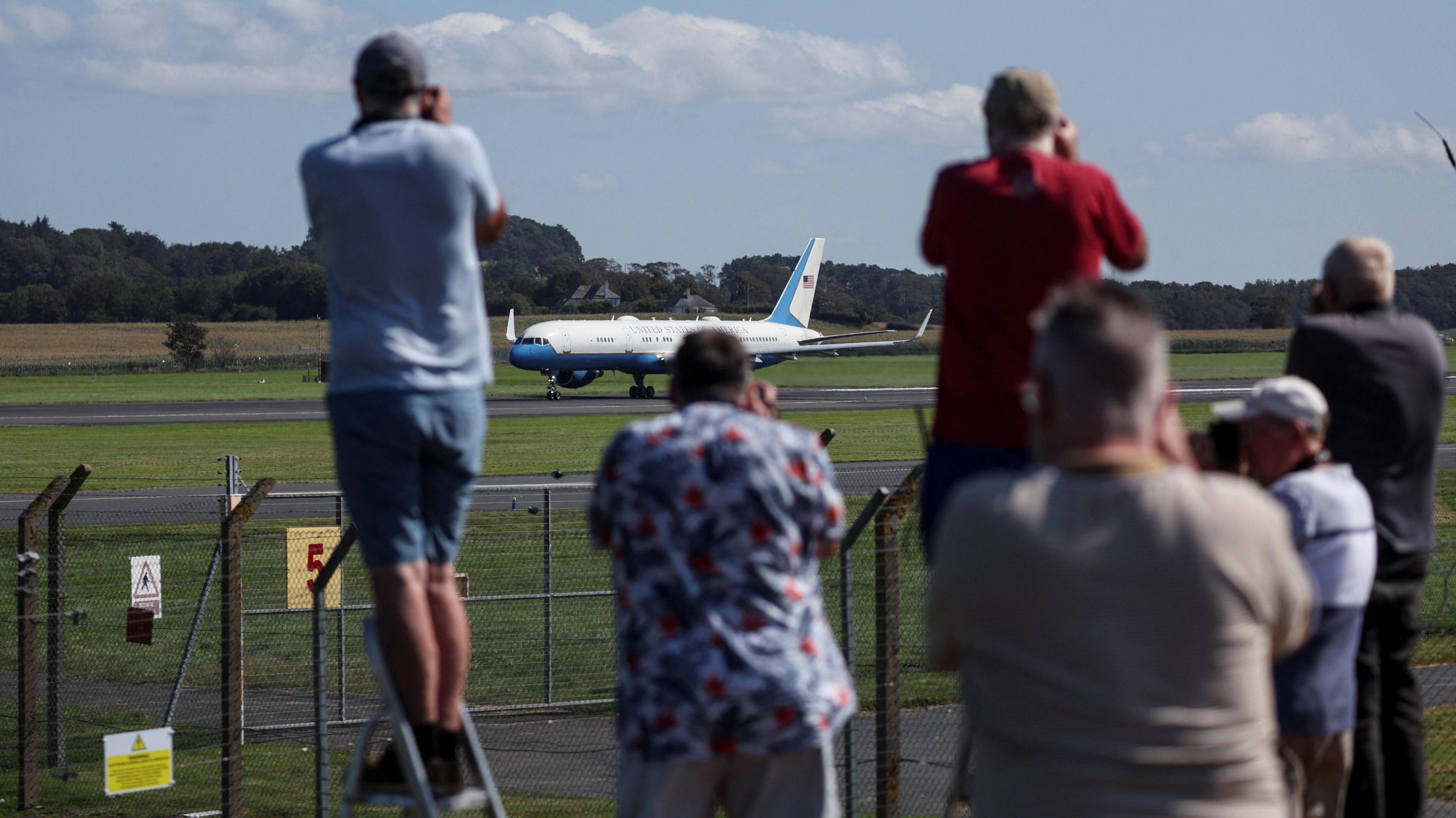 Plane spotters taking pictures of Air Force Two at Prestwick Airport. Two people, one on the left wearing a light blue top and one on the right wearing a red top, are on step ladders. A man in a multi-coloured white, blue and red top is in the middle of the image. They are blurred. The plane in the background is in full focus and is mainly white with blue accents.