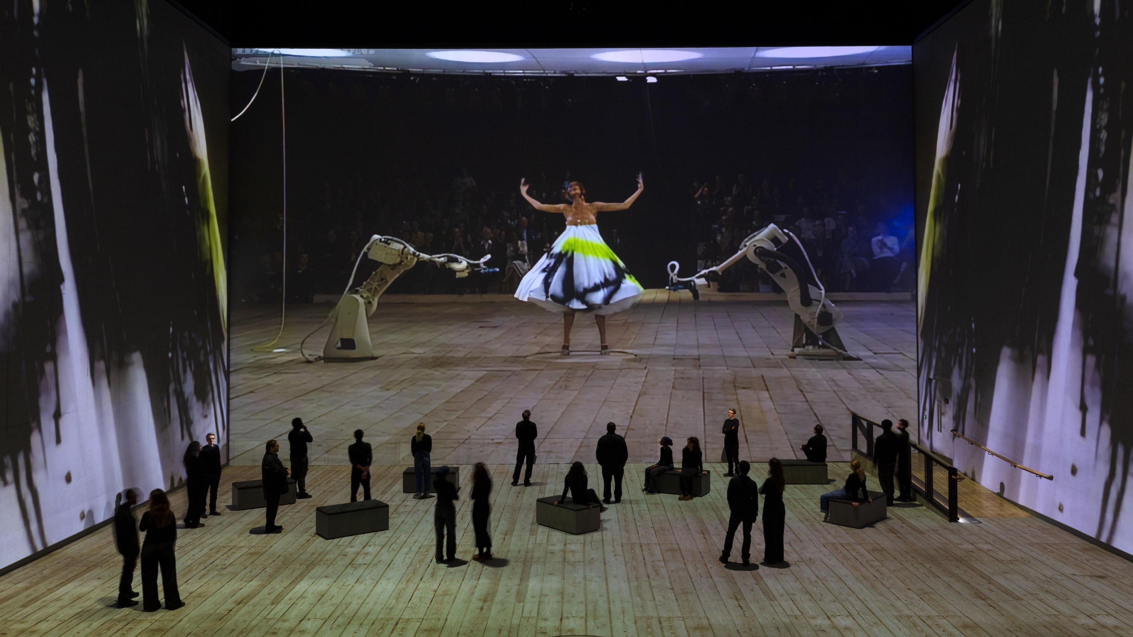 Interior of the Lightroom in London with silhouetted figures in front of a huge screen displaying an image of a model in a dress being spray-painted by robots, from Alexander McQueen's Spring collection 1999.