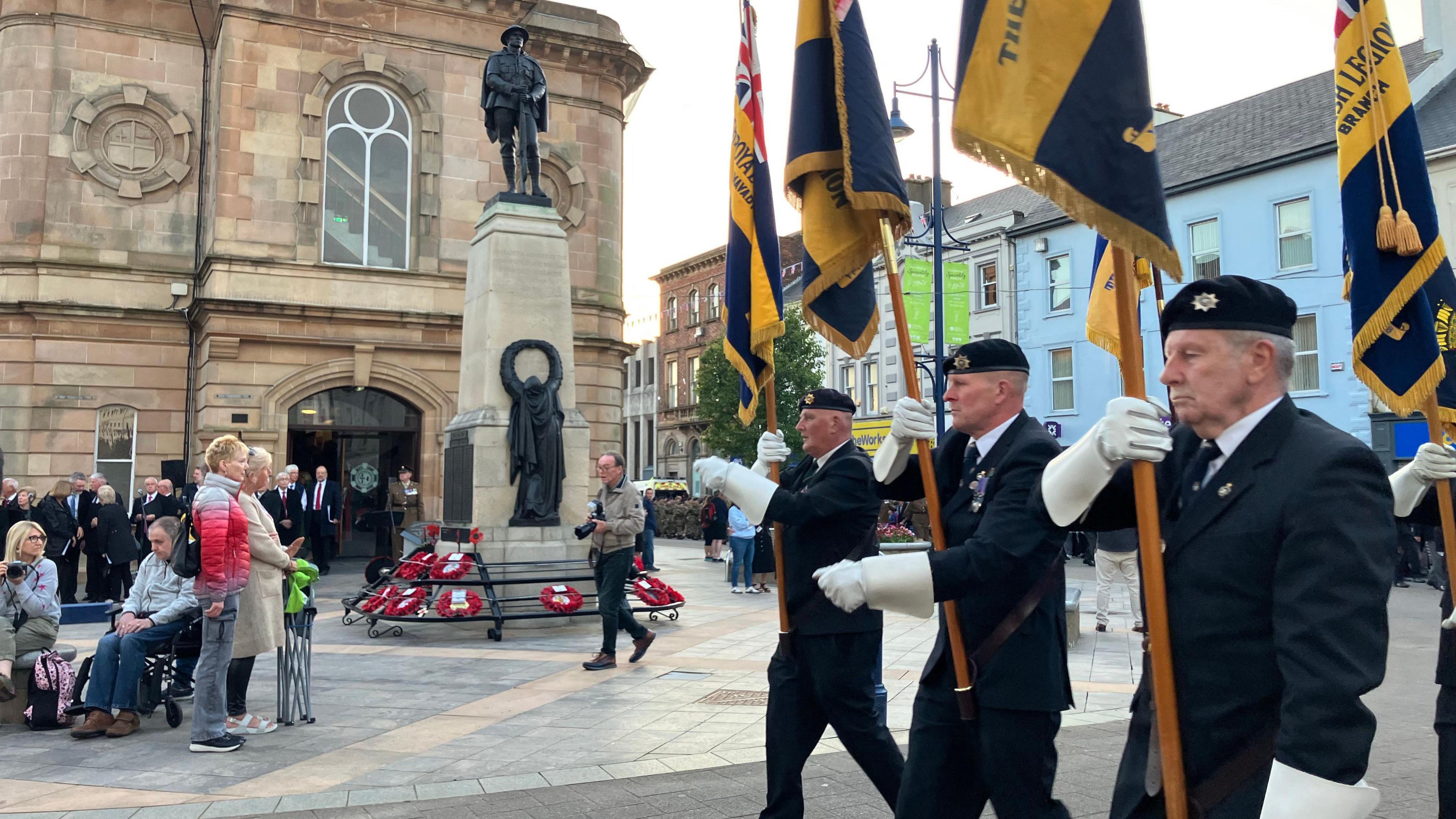 Three men, dressed in navy suits, walking with flags. People are gathered at a statue in the background.