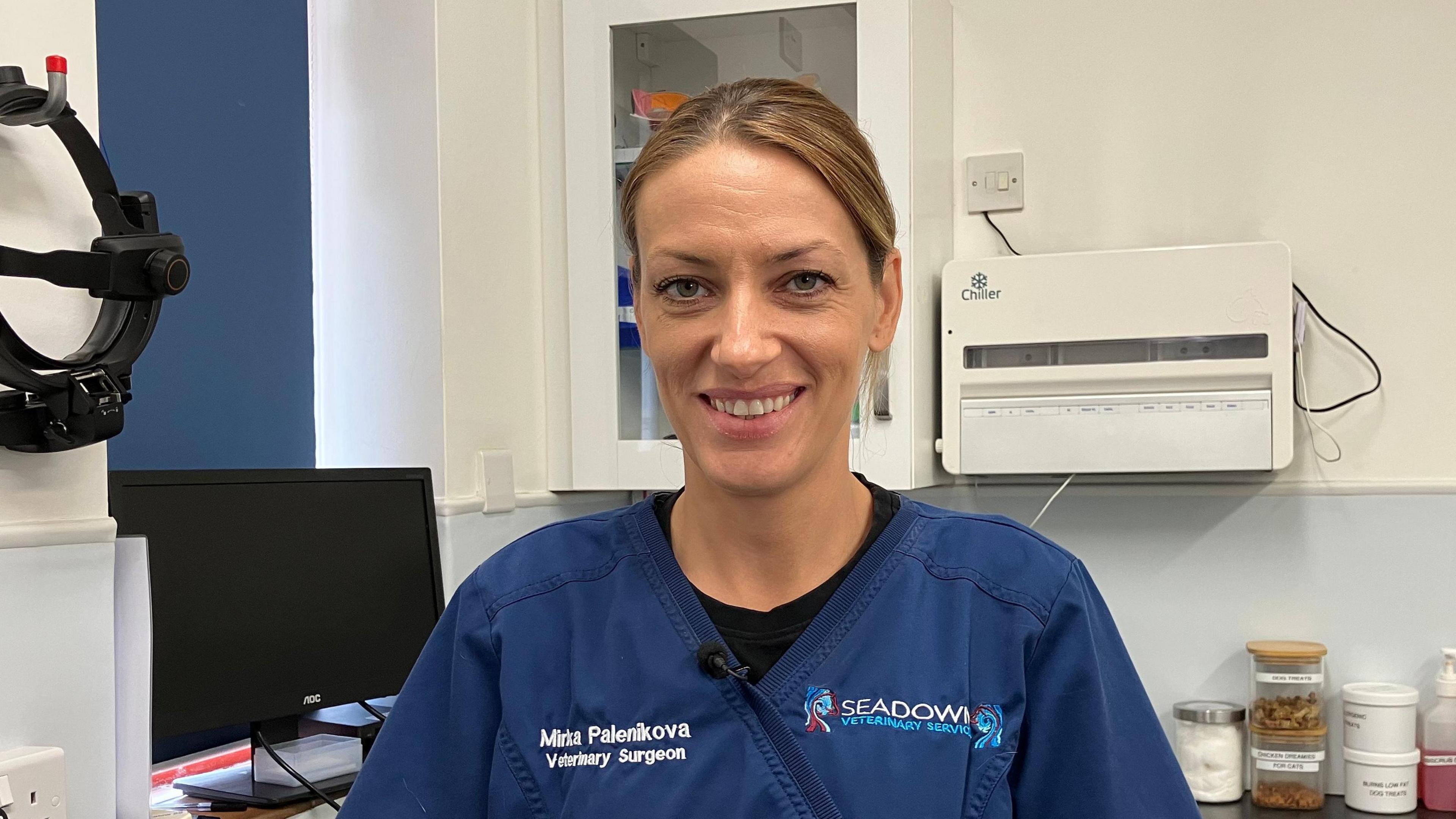 A specialist veterinary ophthalmologist - Mirka Palenikova - is centre frame. She is wearing a blue medical style overall with her name and the name of her surgery on the front, and smiling to camera. She is pictured in the vet surgery with a computer, jars, and equipment in the background.
